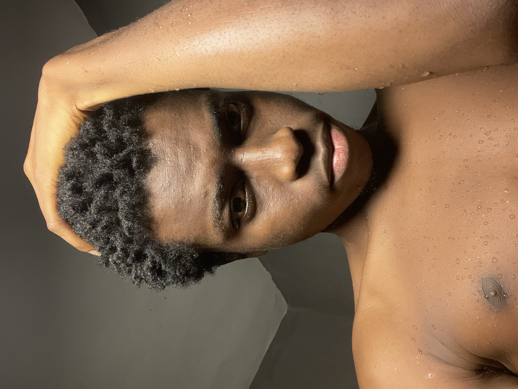 The image is a close-up, selfie-style shot of a young Black male, seemingly in an indoor setting with a gray wall in the background. The subject is shirtless, with water droplets visible on his skin, suggesting he may have recently showered or been in a humid environment. His hand is raised to his dark, curly hair. His expression is serious, with his gaze directed towards the camera. The lighting is dim, creating strong shadows and highlighting the texture of his skin and hair. The location is identified as Nkpor, Nigeria, though no specific landmarks or environmental details within the city are depicted.