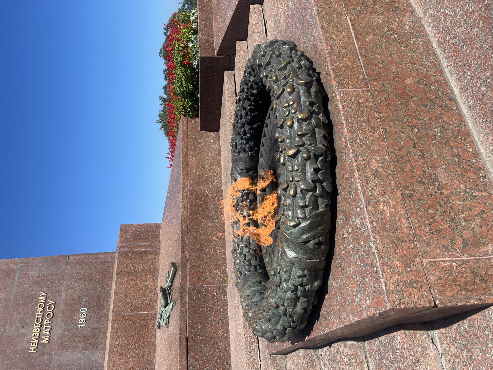 The image shows an eternal flame in a bronze wreath in front of a granite monument in Odesa, Ukraine. The monument has an inscription in Russian that reads "TO THE UNKNOWN SAILOR" and the year "1960". To the left of the wreath, there is a bronze sculpture of a rifle and a helmet. In the background, there are red flowers and a clear blue sky. The scene is outdoors, under bright sunlight.