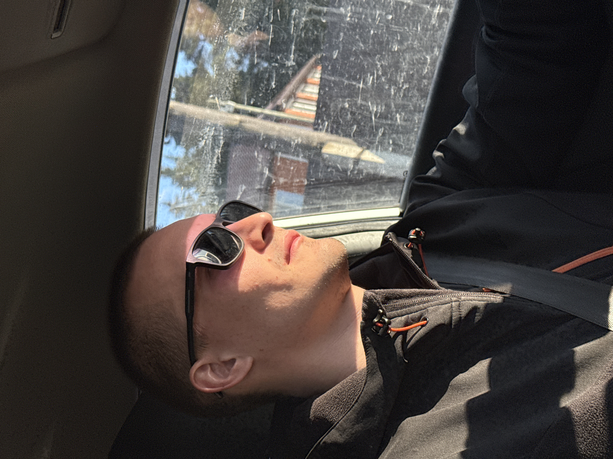 The image depicts a man wearing sunglasses reclining in what appears to be the passenger seat of a car. He is wearing a black jacket with orange accents and a seatbelt is visible across his chest. The car window is grimy and partially obscures the view outside, revealing glimpses of buildings and trees, potentially within an urban area. The presence of the dirty window and the angle of the shot suggest a candid or spontaneous photograph taken within the vehicle. The location is not apparent based on the image.