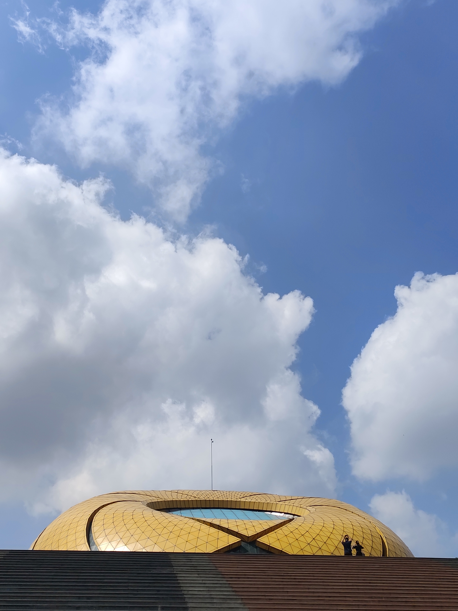 The image shows the rooftop and stairway of the Golden Palace, a popular tourist destination in Da Lat, Vietnam. The rooftop is a golden, dome-like structure with an open-air center filled with blue water. At least two people are visible on the stairs, near the edge of the rooftop. The sky above is partly cloudy, with white cumulus clouds against a blue background. The bottom of the image shows a series of ascending staircases with gray and brown levels.