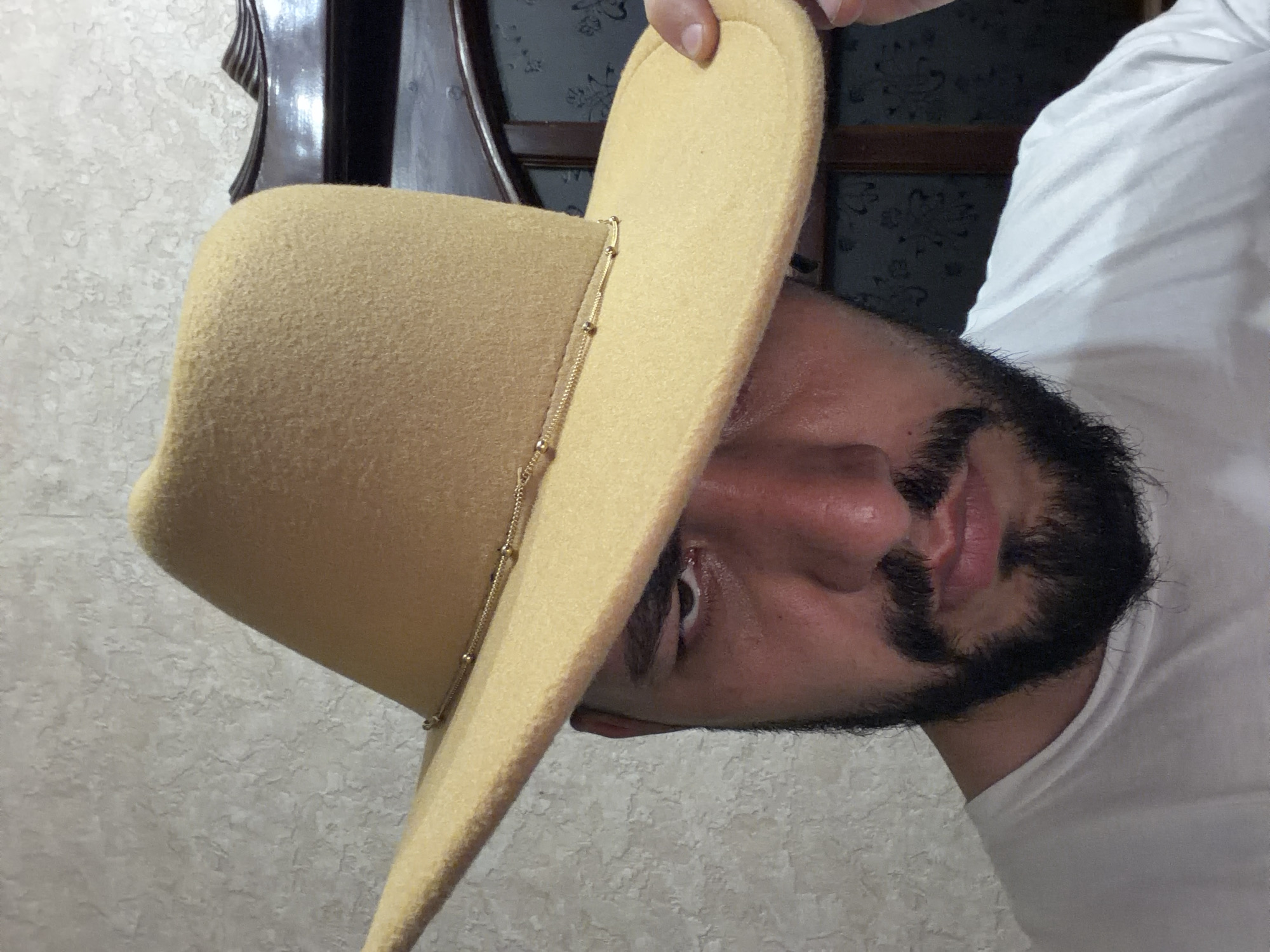 A man with a dark beard and a white t-shirt is holding up a beige cowboy hat. The background features a patterned wall and dark furniture. There is no evidence of Krasnoyarsk, Russia in the provided image.