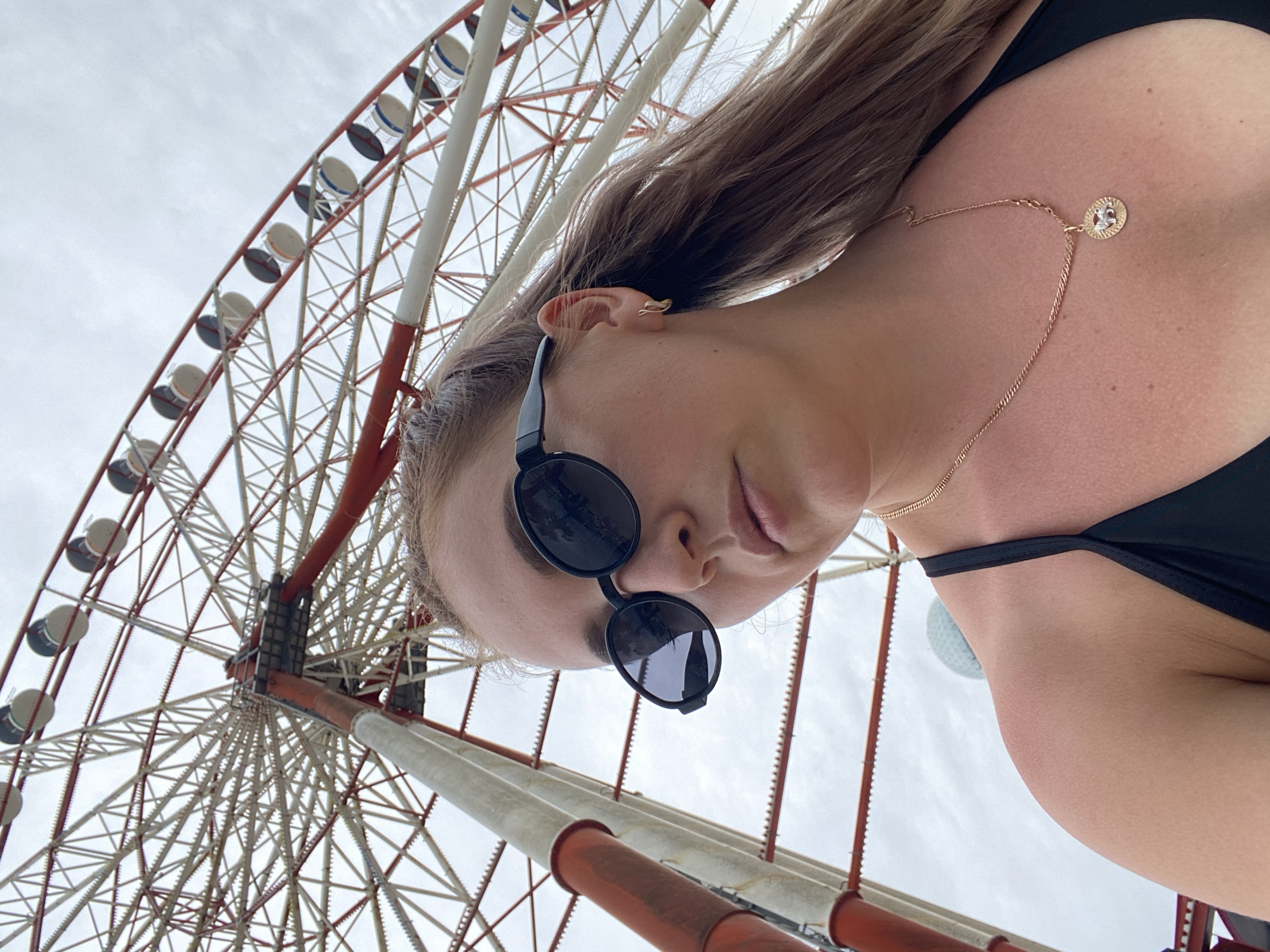 The image shows a young woman with blonde hair tied in a ponytail, wearing black sunglasses, a gold necklace with a pendant, and a black bikini top. She is looking down and to the right. Behind her, a large Ferris wheel with white and red supports is visible against a cloudy sky. The Ferris wheel is a prominent landmark in the city of Batumi, Georgia, located on the coast of the Black Sea. The photo appears to be taken at an amusement park or waterfront area in Batumi.