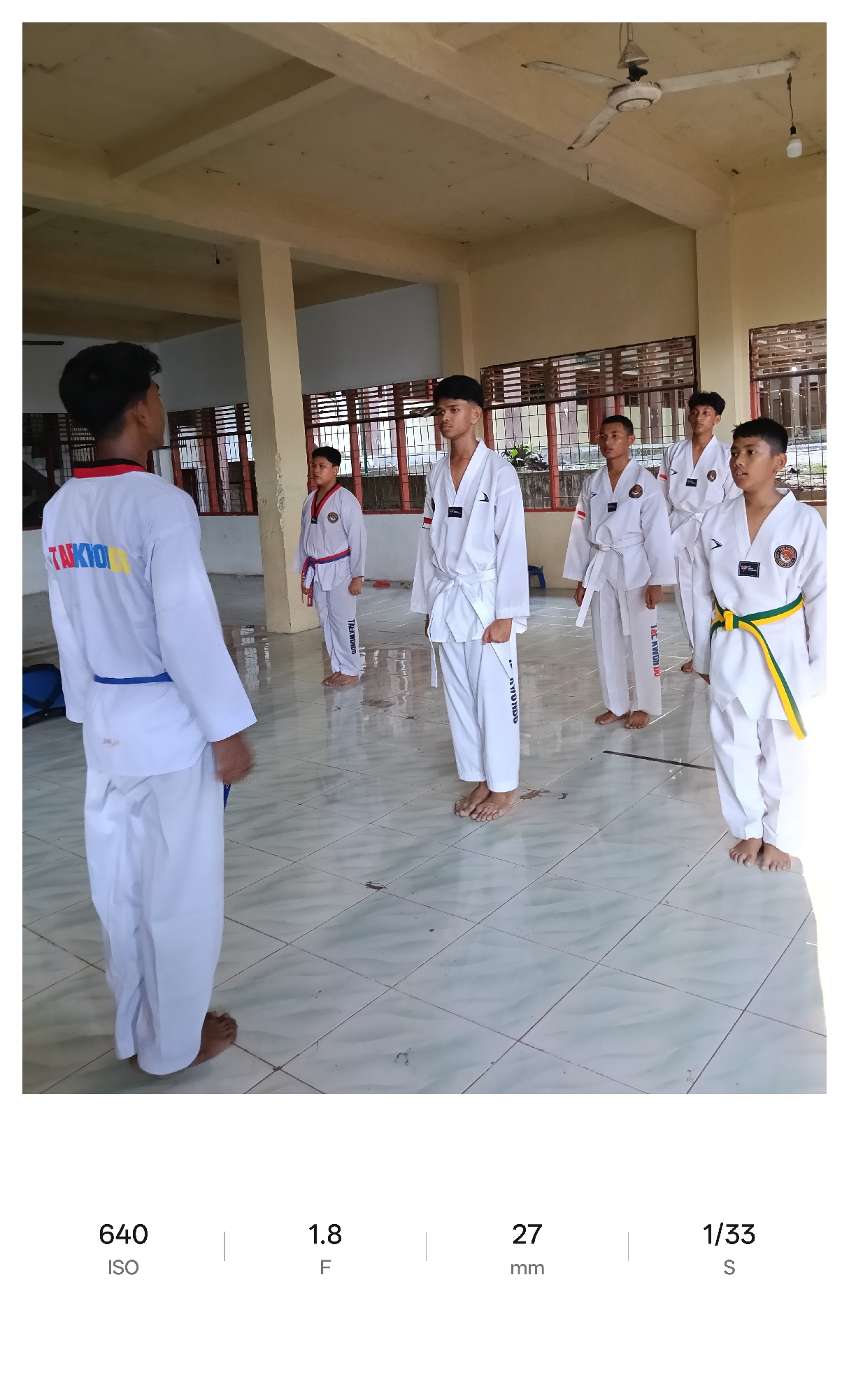 In Lhokseumawe, Indonesia, a group of young males wearing Taekwondo uniforms stand in a tiled room. The uniforms vary by belt color, indicating different ranks, and all individuals are barefoot. One male, with his back to the camera, faces the others. Windows with a red trim are visible in the background. The room is illuminated by overhead lighting, with a ceiling fan also visible.
