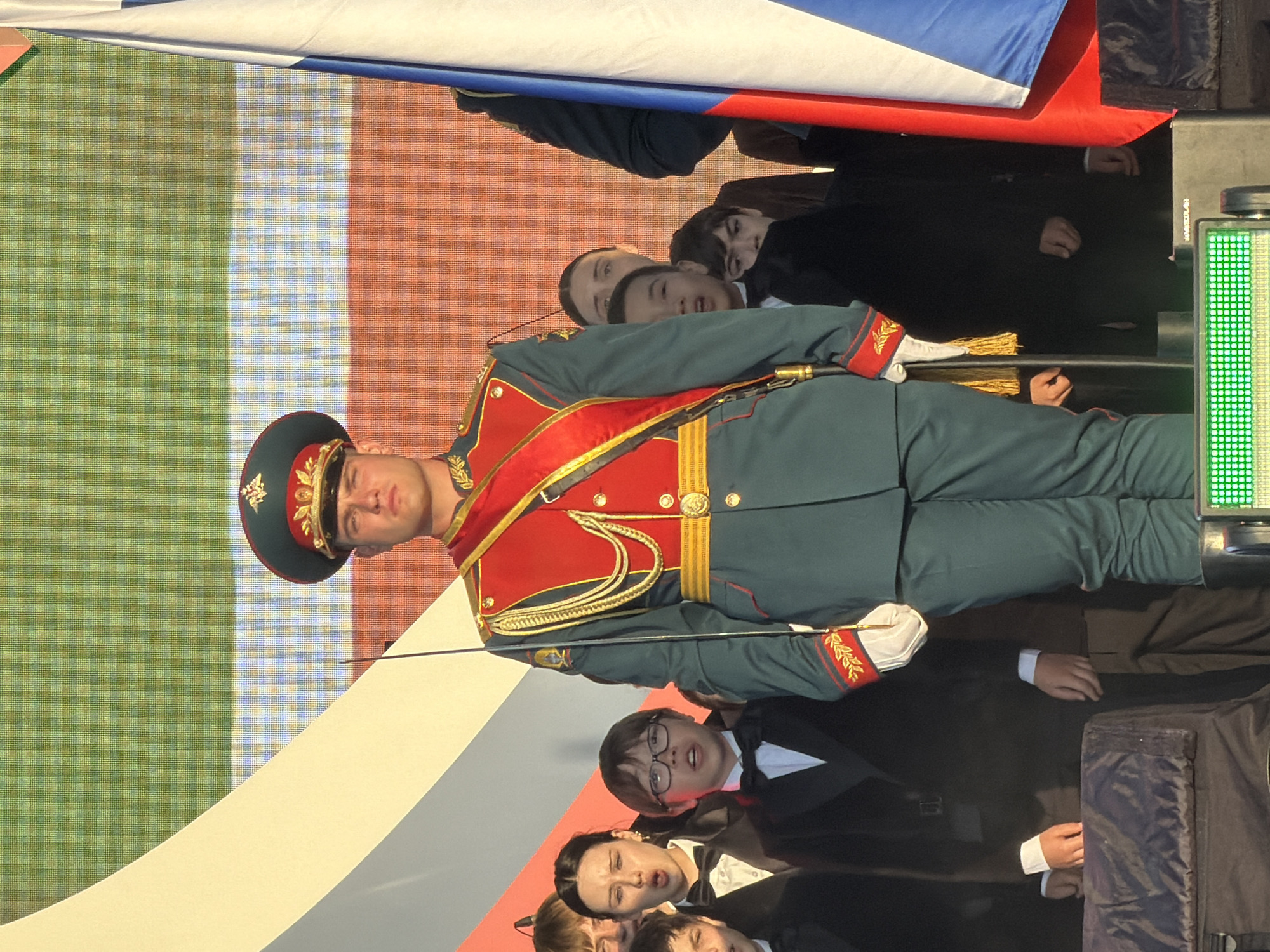 The image captures a formal outdoor event with a central figure in a military-style uniform standing at attention. This individual wears a dark green jacket with red accents, a red sash, and a peaked cap. A sword is held vertically by their side. Behind the central figure, a group of children and young adults are visible, some with their mouths open as if singing or speaking, others looking forward. They are dressed in formal attire, including suits and bow ties. A large flag, appearing to be the Russian flag with its white, blue, and red stripes, is partially visible on the right side of the frame. The background features a large screen displaying abstract patterns and colors, including green, white, and orange. The scene is lit by sunlight, casting a warm glow. The overall setting suggests a patriotic or ceremonial occasion.