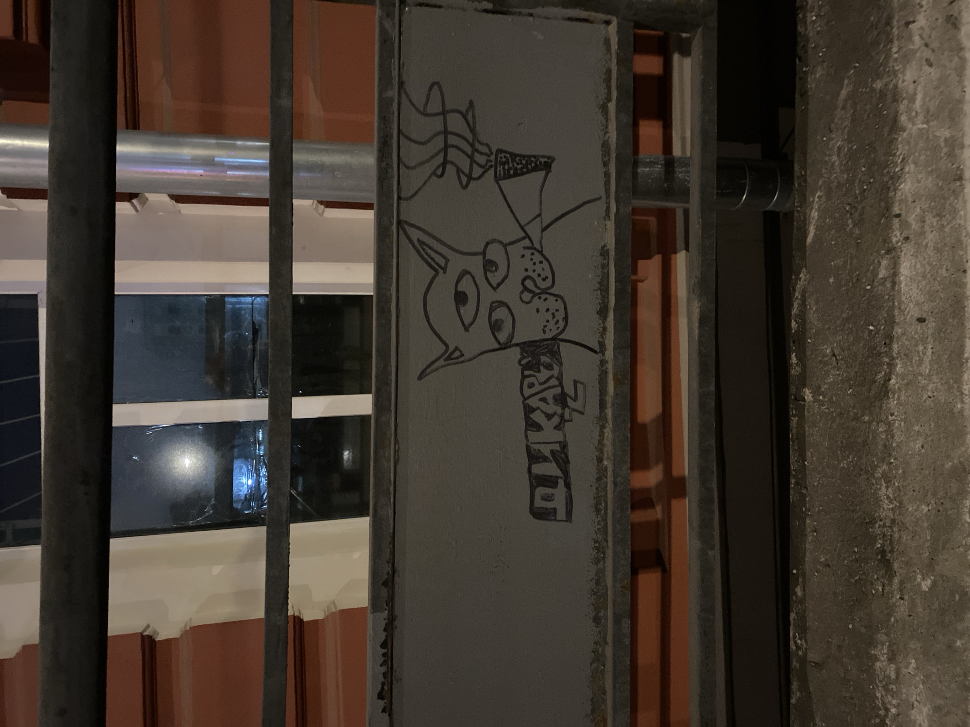 A nighttime view shows a gray metal support beam with a drawing in black ink on its surface. The drawing depicts a stylized dog's head with large, exaggerated eyes and a musical instrument (possibly a trumpet) extending from its mouth, indicated by wavy lines. Below the dog is the inscription "ДЖКАРВЪ" in a vertical arrangement. The metal support beam is part of a metal grate or fence structure, which is positioned in front of a building. Visible through the grate are window frames with white trim against a reddish-brown building facade. The right side of the image reveals a concrete wall. The location is assumed to be in Moscow, Russia based on the Cyrillic lettering.