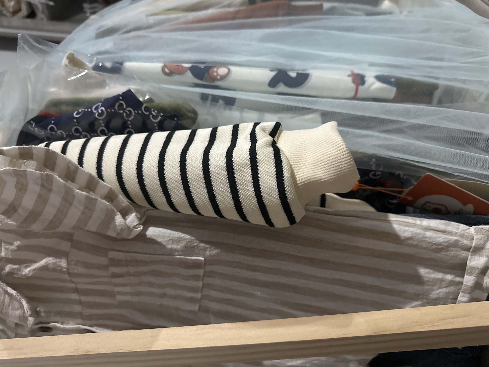 A close-up shot shows a rack of children's clothing. The main garment in focus is a beige and white striped shirt with a ruffled collar and sleeves. To the right, a white sleeve with black stripes is visible, as is a light blue tulle fabric. Behind these items, other garments are partially visible, including one with a pattern of interlocked Gs and another with a teddy bear print. The background is out of focus and suggests an indoor setting, possibly a closet or boutique. The image does not provide any information about its location in Тайгараев, Kyrgyzstan.