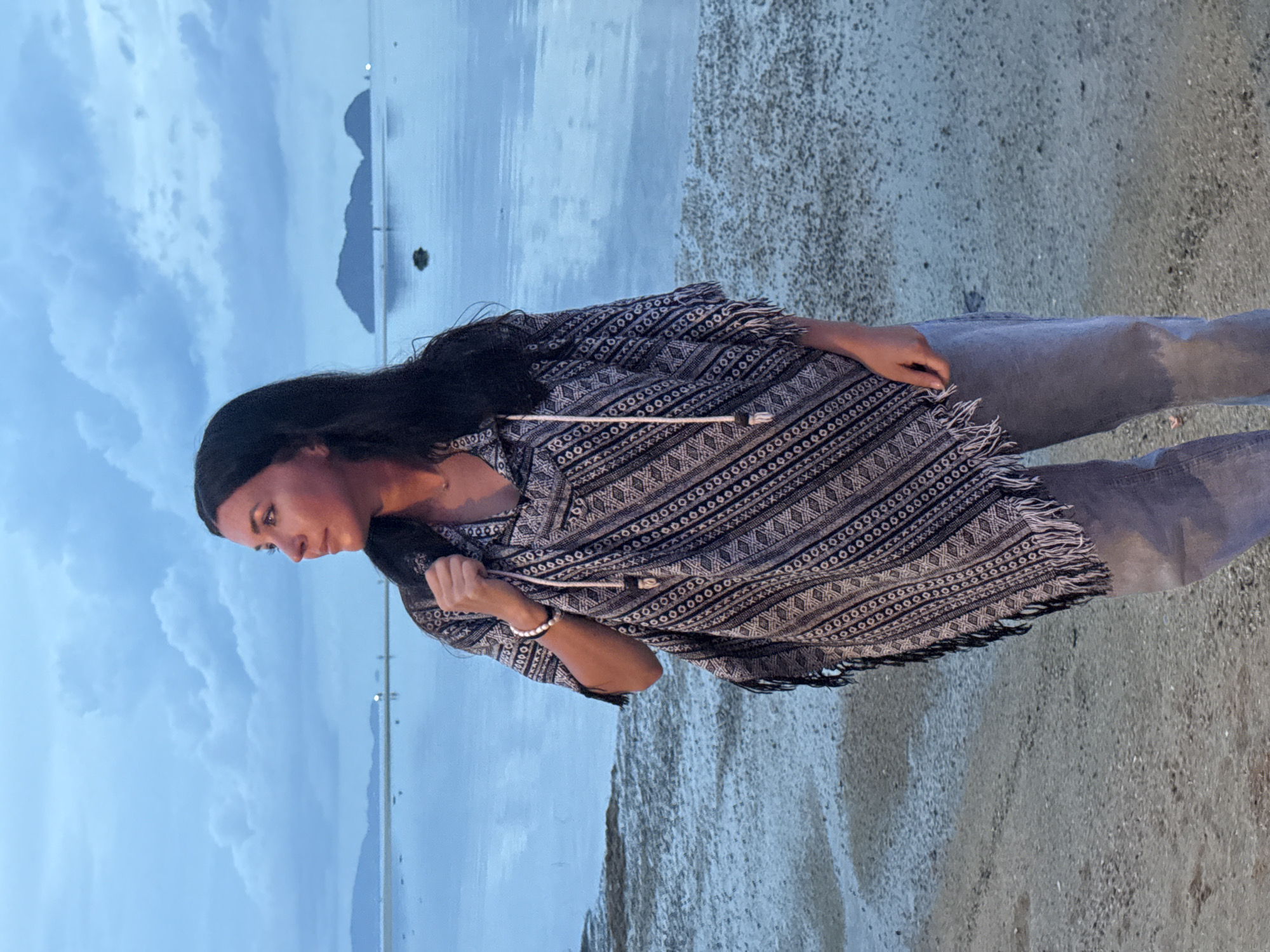 A young woman stands on the shore in Baan Sri Thanu, Thailand. She is wearing grey jeans and a patterned black and white poncho-style top with fringe. She has long dark hair and a black headband. She looks down and to the left. The ocean stretches out behind her, and in the distance, the silhouettes of mountains are visible under a cloudy sky. The ground is sandy.