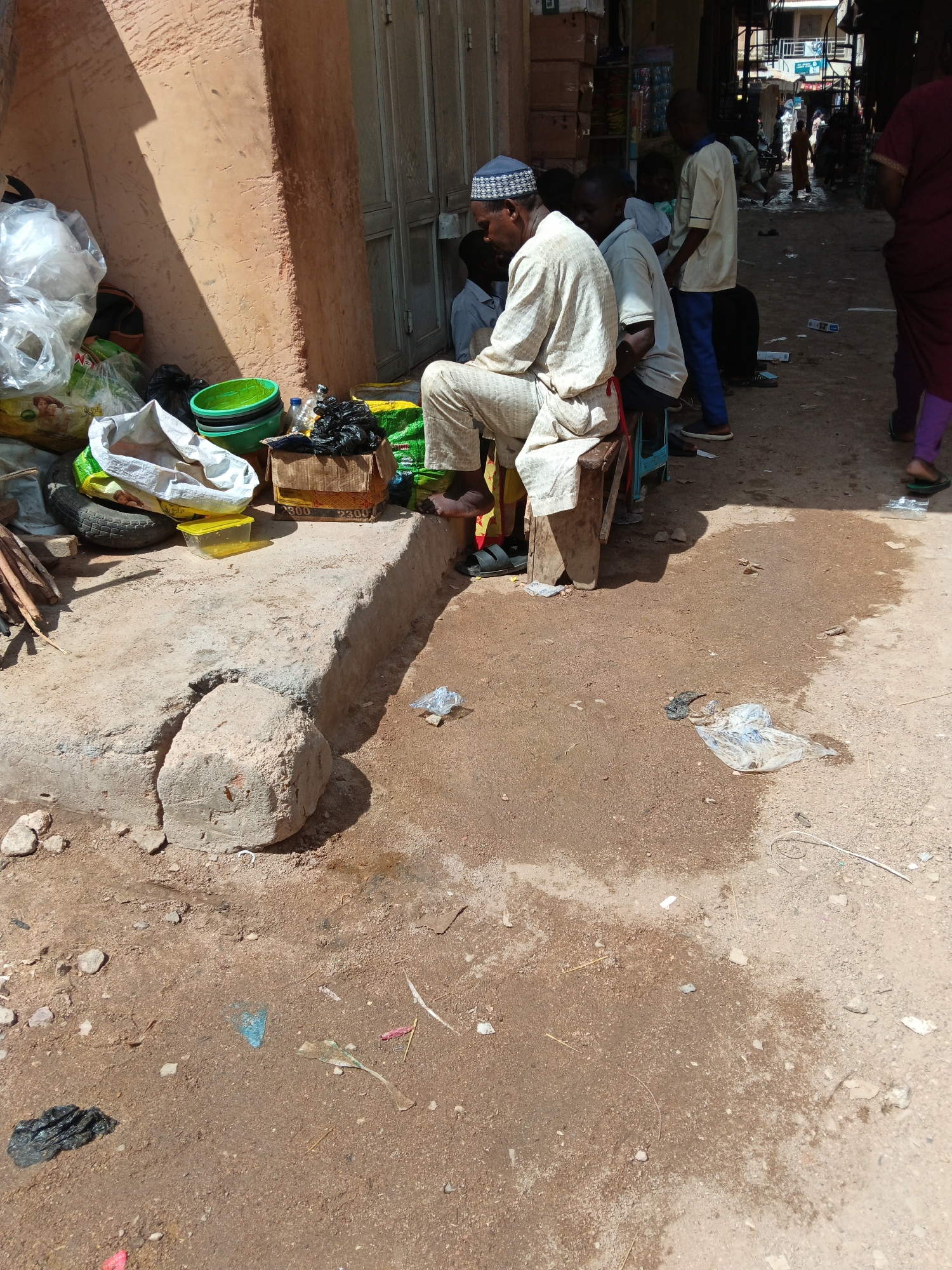 In Arawa, Nigeria, a street scene unfolds under the midday sun. A group of individuals, primarily men and boys, are gathered near a building with an orange-toned, textured wall and a large, light-colored door. The ground is a mixture of dry, dusty earth and some damp patches.
In the foreground, a man in a light-colored robe and a patterned cap sits on a wooden stool, facing away from the camera. He appears to be observing something to his right. To his left, a collection of items is piled near the wall. These include several large, white plastic bags, a green plastic basin containing smaller green basins, a yellow plastic container, a tire, and several bundles of sticks. A brown cardboard box, with some writing on it, sits on a raised concrete ledge, next to a pile of black plastic bags.
Further back, other individuals are visible, some standing and some sitting. One young boy in a light-colored shirt and blue pants stands facing left, while another person in a dark garment and purple leggings walks away from the camera on the right side of the scene. The background suggests a market or street environment with more buildings and structures, including what appear to be shop stalls. Litter, such as plastic wrappers and scraps of paper, is scattered on the ground. The overall impression is one of everyday life unfolding in an outdoor, possibly commercial, setting.