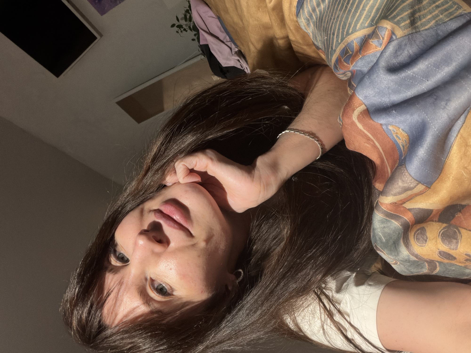 This indoor image, set in Perm, Russia, features a woman lying down and looking towards the camera. Her head is tilted, giving a low-angle perspective. She has long, dark brown hair that cascades around her. One hand is gently placed near her chin, drawing attention to her face.
The woman's expression appears calm and neutral, with her blue-grey eyes directly engaging the viewer. Her lips are slightly parted. She is wearing a silver chain-link bracelet on her left wrist and a small hoop earring in her right ear. A white top is visible.
A significant portion of the image is filled by a colorful, patterned blanket or scarf, featuring geometric and abstract designs in tones of blue, orange, yellow, and beige. The background shows a ceiling with two dark, rectangular objects mounted, possibly framed pictures or panels, and a glimpse of a green plant in the upper left. The lighting is distinct, suggesting an evening or nighttime setting, with a strong light source illuminating parts of her face and creating pronounced shadows. There is no visible text in the image.