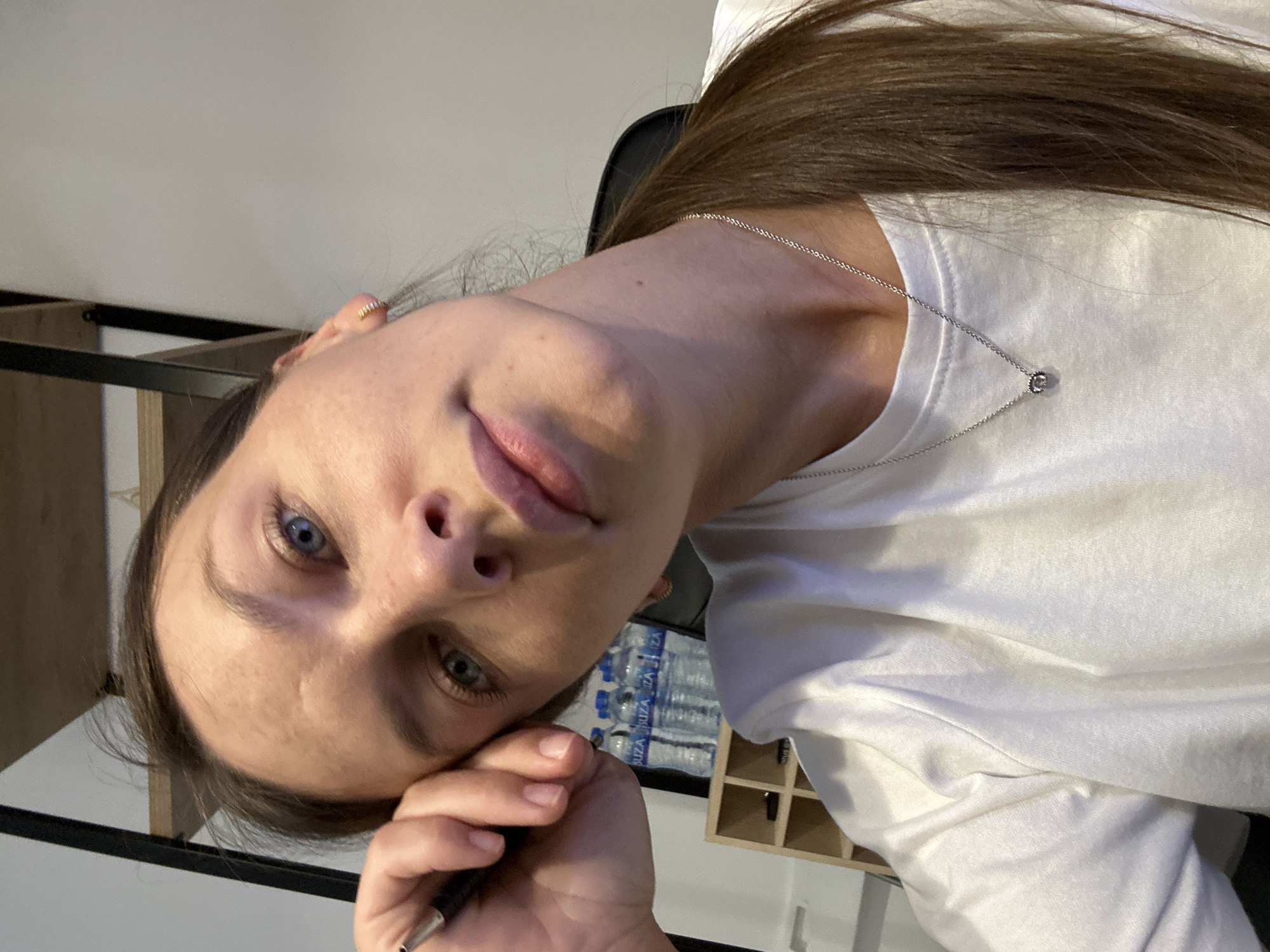 A woman is seen in a close-up shot, leaning her head on her hand, which is holding a pen. She has blue eyes, light skin, and brown hair pulled back in a ponytail. She is wearing a white t-shirt and a silver necklace with a small circular pendant. In the background, there are several bottles of water and a wooden shelf. The scene appears to be indoors, possibly an office or a home. There are no overt indications of the specific location being Boreti, Montenegro, beyond what is requested to be inferred.
