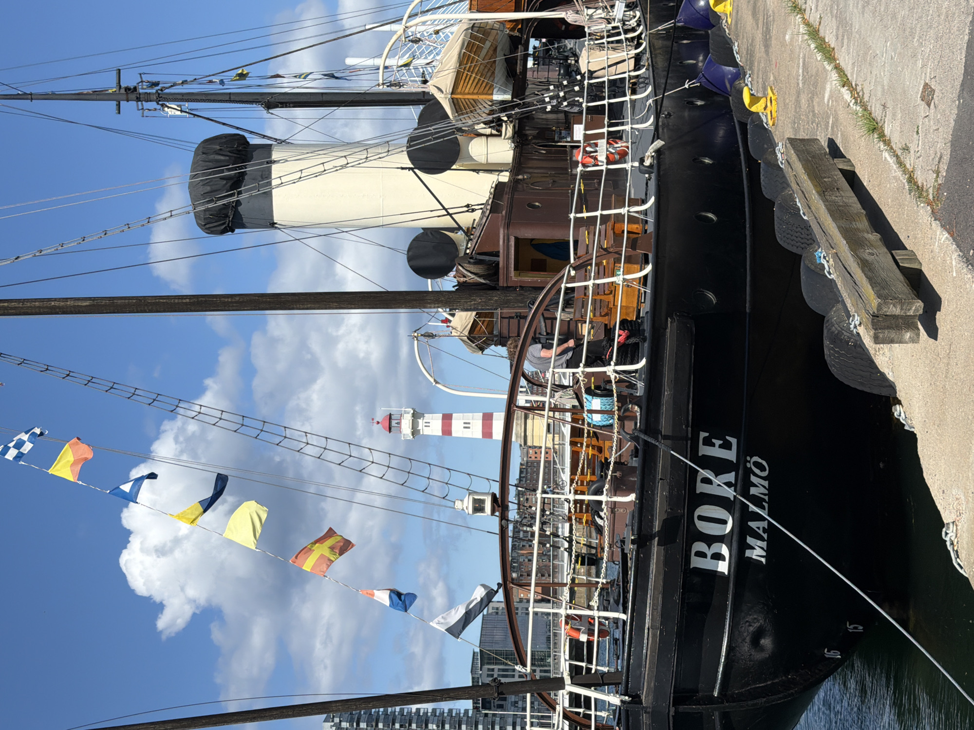 The image shows a black ship named "BORE MALMÖ" docked at a harbor in Malmö, Sweden. The ship has a tall smokestack covered with a black tarp and is adorned with various international maritime signal flags. In the background, a red and white striped lighthouse stands tall. The ship's deck is visible with railings and a life preserver. A person in a grey t-shirt is seen standing on the deck. The harbor area is paved with concrete and has tires and yellow bollards along the edge of the dock. The sky is blue with scattered white clouds, suggesting a clear day.
