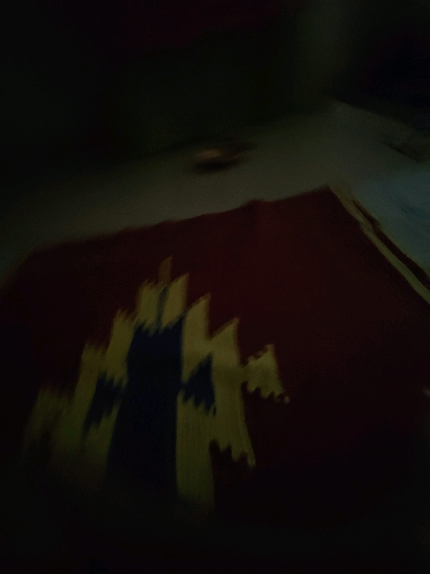A blurry, low-light image shows a dark maroon fabric with a geometric pattern in yellow and dark blue. The pattern resembles stylized representations of buildings or mountains, with pointed peaks and a central dark blue shape that could be interpreted as a figure or structure. The fabric appears to be a textile, possibly a rug or tapestry. The surrounding environment is very dark, with some faint, indistinct shapes visible in the background, suggesting an indoor setting at night or in low light. There are no people or discernible actions depicted in the image. The location within Pune, India, cannot be determined from the visual information provided.