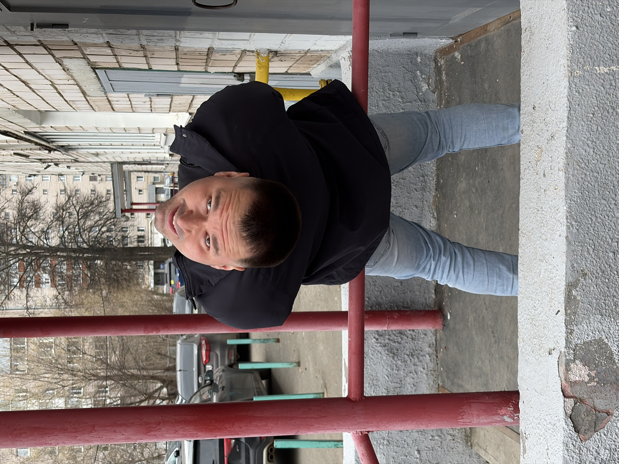 The image shows a man standing outside a building, possibly in Izhevsk, Russia. He is wearing a black jacket and light-blue jeans. He is located behind a red metal railing. Behind the railing, there are parked cars and bare trees, and a residential building is visible in the background. The ground is concrete, and there is a white concrete curb or step on the right side of the frame. The building in the foreground appears to be constructed with white bricks.