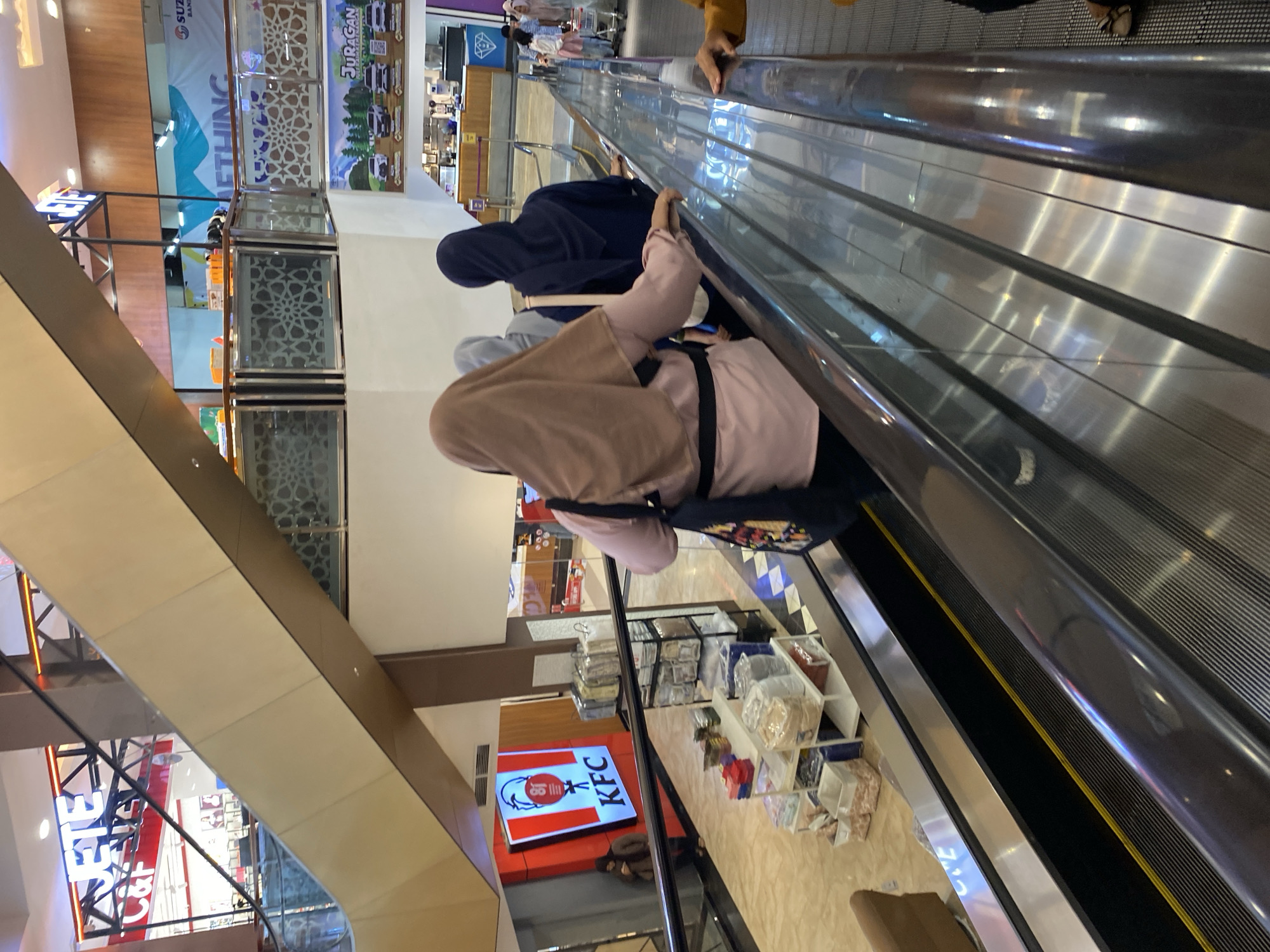 The image depicts a shopping mall in Banda Aceh, Indonesia, with a focus on an escalator. Several individuals, predominantly women wearing hijabs, are visible on the escalator. One woman in a light pink shirt and beige hijab is in the foreground, facing away from the camera and holding onto the escalator handrail. Another woman in a dark blue hijab is positioned behind her, also facing away. In the background, a prominent KFC sign is displayed on a red illuminated panel. Various retail displays are present, including shelves filled with boxed items and a display of bedding. The overall environment suggests a commercial setting, likely a multi-story shopping complex with escalators connecting different levels. Advertisements for businesses like "JURAGAN" and "SUZUKI" are also visible, further confirming the mall's commercial nature. The lighting is typical of an indoor retail space, with artificial lights illuminating the scene.