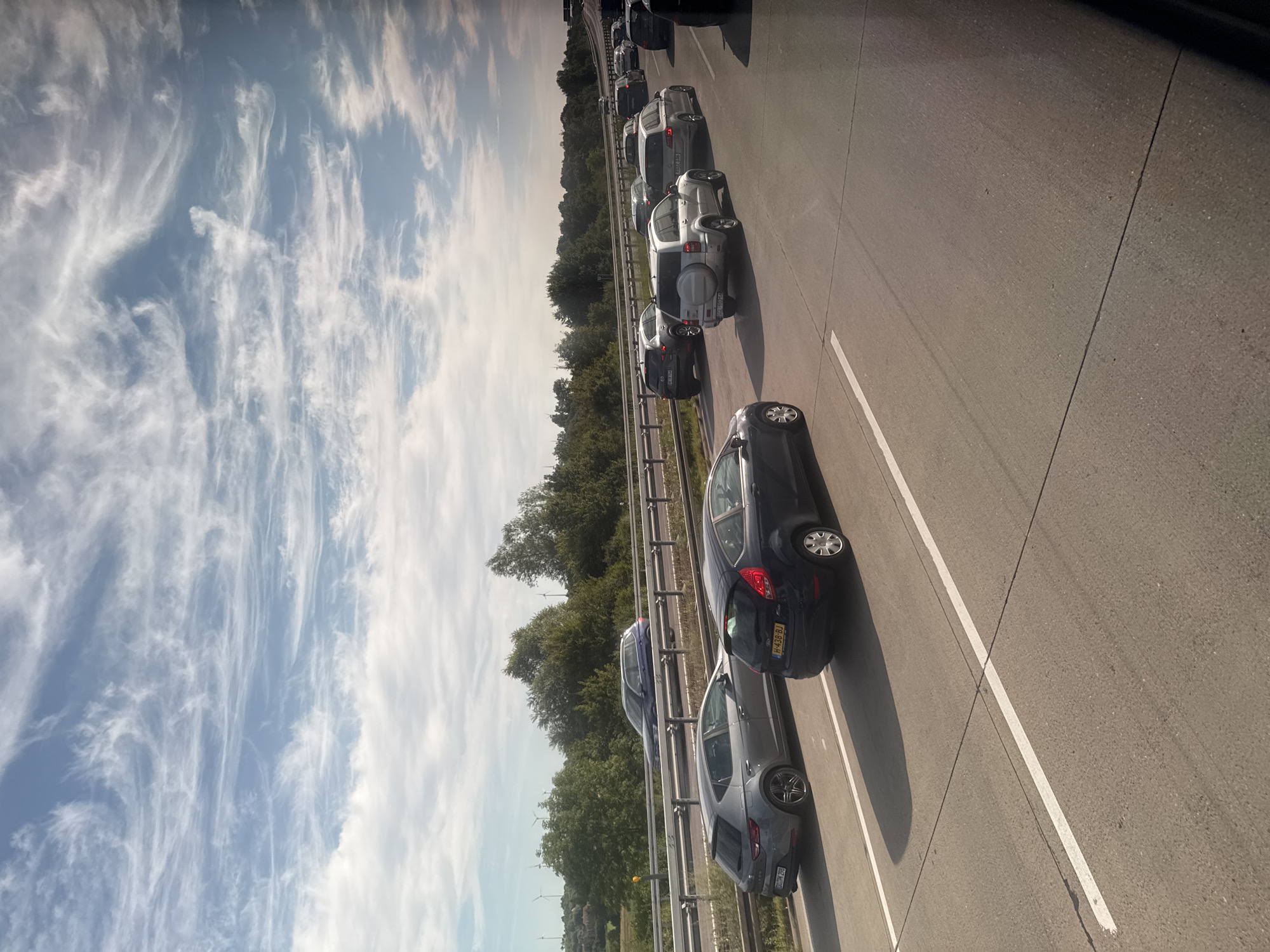 The image shows a traffic jam on a highway. Several cars are visible, including a dark blue Ford Fiesta, a grey Seat Leon, and a silver SUV. The cars are driving on a paved road with white lane markings. In the background, there are trees and a blue sky with wispy clouds. The scene appears to be in a rural area, with wind turbines visible in the distance. The time of day is daytime, as indicated by the bright sky and shadows.