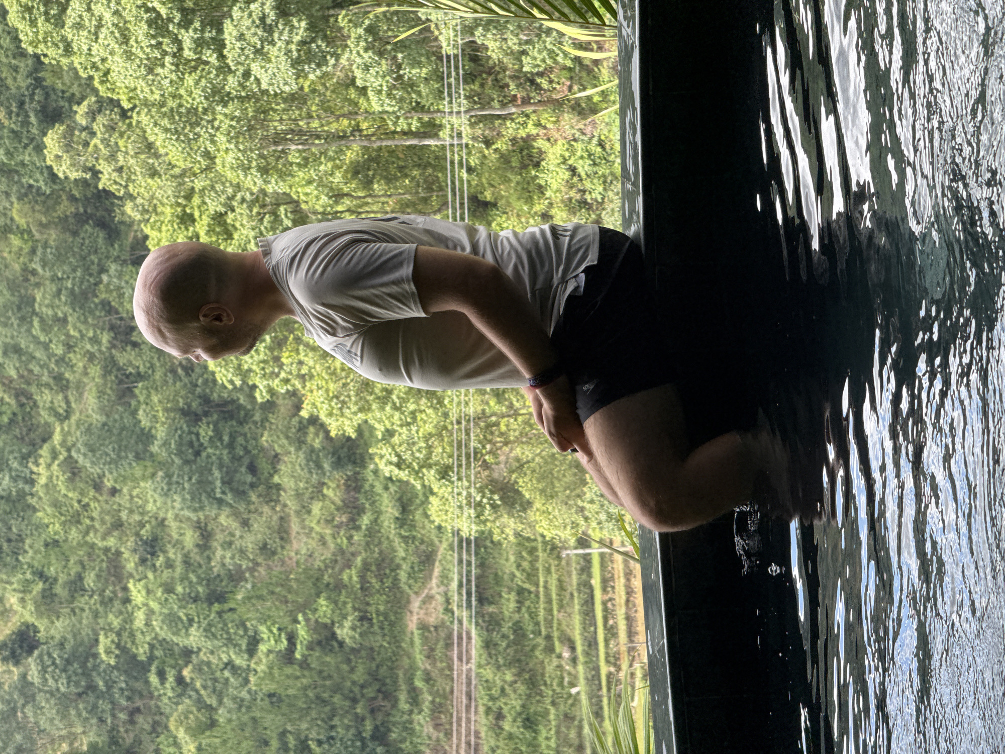 A bald man, viewed from the side, sits on a dark stone ledge with his lower legs and feet submerged in rippling water, facing left towards a verdant mountainous landscape. He wears a light grey short-sleeved t-shirt and dark shorts, with a multi-colored bracelet on his left wrist. The water reflects shimmering light, indicating an outdoor setting, likely an infinity pool or natural spring. In the background, the scene reveals lush green, densely forested mountains, characteristic of the Mù Cang Chải region in Vietnam. Several tiers of agricultural terraces, consistent with rice paddies, are carved into the lower slopes of the mountains. Three parallel utility lines are visible horizontally across the midground of the landscape. The overall impression is one of a man relaxing and contemplating the natural, agricultural scenery.