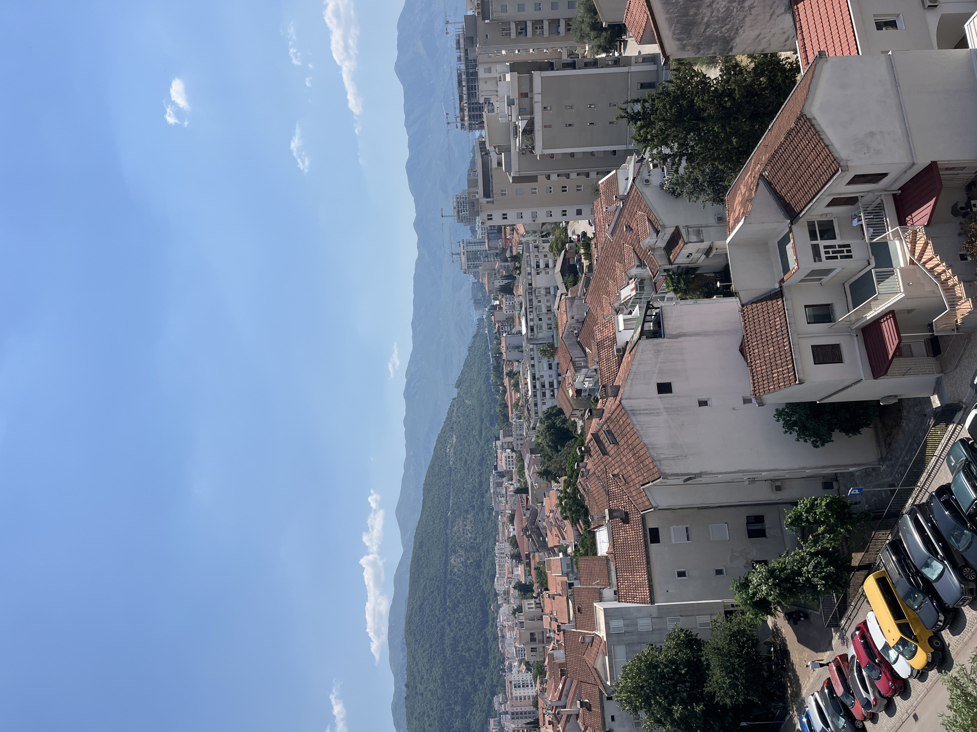 The image depicts an elevated, wide-angle view of a city, likely Budva, Montenegro, characterized by its dense urban development and mountainous backdrop. In the foreground, numerous buildings with terracotta tile roofs are visible, interspersed with residential apartments and smaller structures. A parking lot in the lower left corner shows a variety of cars, including a yellow minivan, a white hatchback, a red hatchback, and several dark-colored SUVs. A single individual, wearing dark pants and a white shirt, is walking away from the parked cars.
The middle ground reveals a more extensive cityscape with taller apartment buildings and ongoing construction, indicated by cranes and unfinished structures. The urban sprawl extends towards a lush, green mountainous region that dominates the background. The sky is a clear, bright blue with a few scattered white clouds. The overall impression is a sunny day in a coastal city with significant development and a prominent natural landscape.