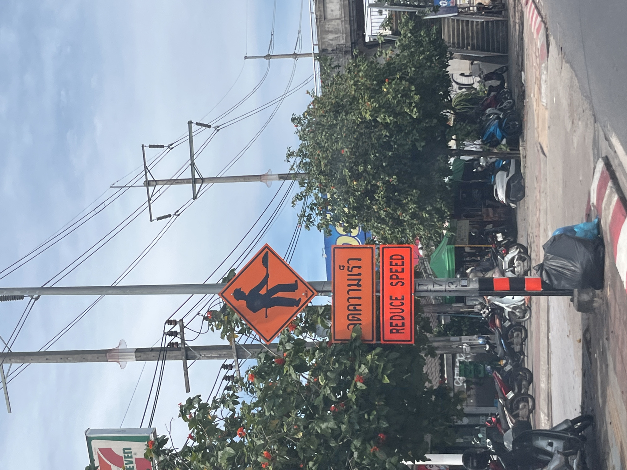 A street scene in Rawai, Thailand, features a traffic pole with multiple signs. The topmost sign is diamond-shaped and orange, depicting a black silhouette of a person shoveling. Below this is a rectangular orange sign with Thai text, followed by another rectangular orange sign with the English text "REDUCE SPEED." The pole itself is gray and black. To the left of the pole is a lush green tree with red flowers, and a 7-Eleven store sign is partially visible in the background. To the right, a tree with denser foliage partially obscures a concrete overpass and a set of stairs. Parked motorcycles are visible on the road and sidewalk, and a black trash bag lies on the ground near the base of the traffic pole. The sky is cloudy.
