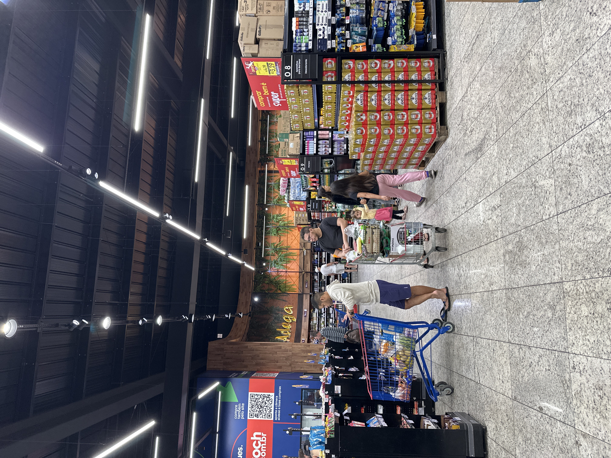 The image depicts the interior of a grocery store, specifically focusing on a section with beer and other goods. Overhead are linear lights, and the store features a dark, modern design. Several customers are present, each pushing a shopping cart filled with groceries. The shelves are stocked with various products, including beer cans arranged in stacks, deodorants, shaving products, and other miscellaneous items. A large sign above the shelves reads "Adega," and another display with a QR code invites customers to shop online. The floor has a speckled pattern. The image was taken at Koch supermarket in Florianópolis, Brazil.