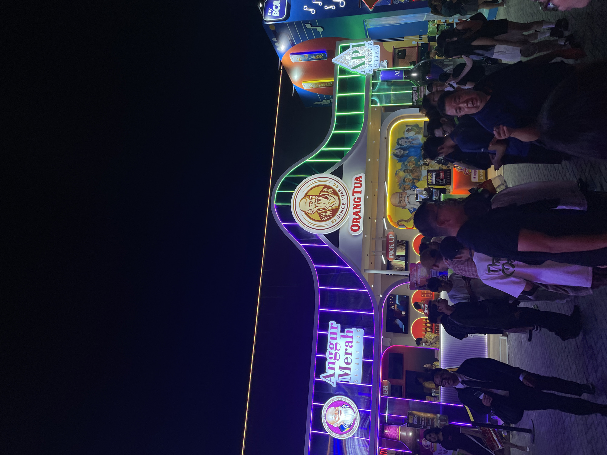 The image shows an outdoor nighttime scene in Salembaran, Indonesia, likely at a public event or fair. A large, brightly lit archway serves as a focal point, adorned with the "Orang Tua" and "Anggur Merah" logos. The archway also displays neon lights, and the surrounding structures have similar colorful lighting. A crowd of people is gathered near the structure. Some of them are looking at the camera. The ground is paved with light-colored bricks or tiles. The background is mostly dark.