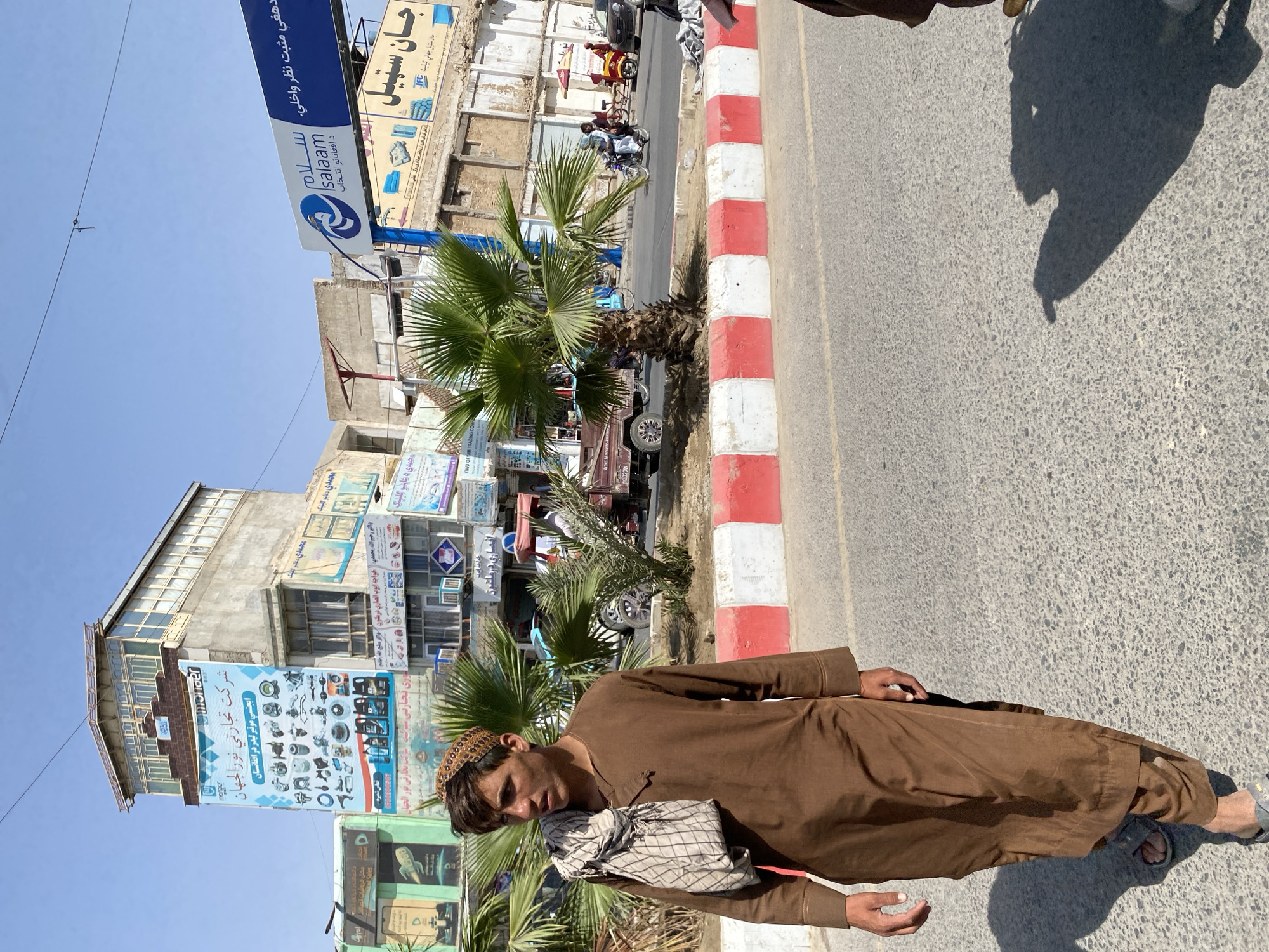 A young man in a brown shalwar kameez and a patterned skullcap walks on a paved street in Kandahar, Afghanistan. He carries a patterned scarf draped over his right shoulder. Behind him, a multi-story building displays advertisements in Arabic and English for various products, including automotive parts and oil. A blue and white sign with the word "salam" is visible above other storefronts. Palm trees line the street, and a red and white striped barrier separates the sidewalk from the road. Several vehicles, including a three-wheeled truck and motorcycles, are present on the street, suggesting it is a busy commercial area. The sky is clear and blue.