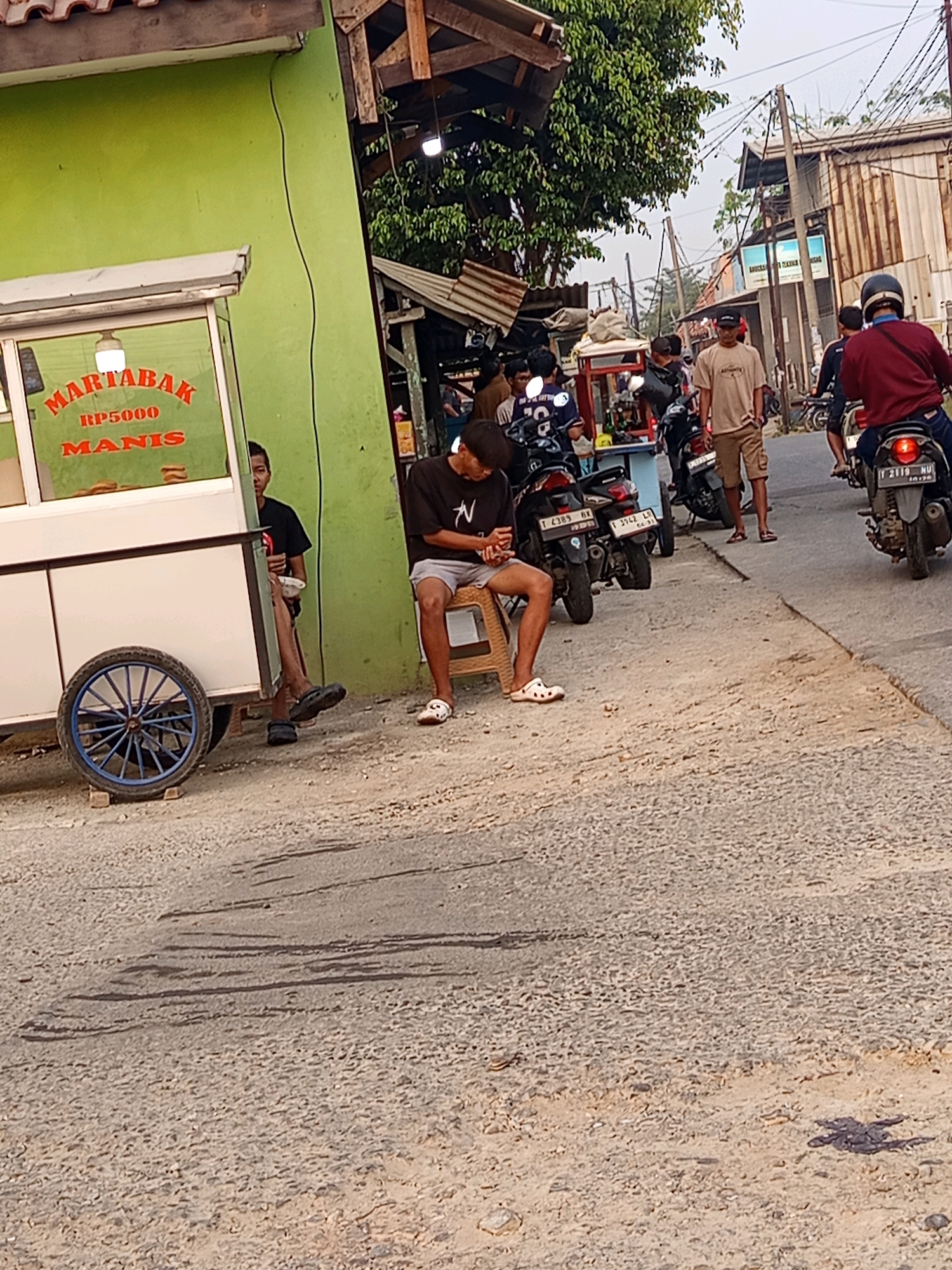 The image depicts a street scene in Karawang, Indonesia, likely a roadside market or food stall area. In the foreground, a green building with a white food cart stands to the left. The cart has text indicating "MARTABAK" and "RP5000 MANIS". A person is seated on a stool next to the cart, looking down. Further down the street, several motorbikes are parked and some people are visible. One person is riding a motorbike with a red jacket. The street is paved with asphalt and gravel, and there are utility poles and wires overhead. The overall atmosphere suggests a bustling, everyday scene in an Indonesian city.