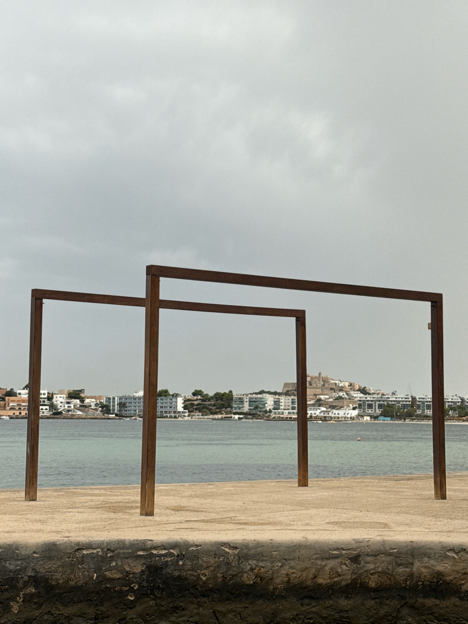 The image depicts an outdoor scene with a body of water in the mid-ground, beyond which lies a cityscape on a slight elevation. In the foreground, there is a concrete surface with a rough, dark stone-like base at the very bottom. Dominating the foreground are two modern, rust-colored metal rectangular frames, positioned close to each other. The larger frame is on the right and consists of two vertical posts and a horizontal beam. The smaller frame is to the left and appears to be of similar construction. The water is calm and has a greenish-blue hue. The cityscape comprises numerous white buildings with some colored rooftops, and trees. A prominent structure, possibly a castle or cathedral, sits atop the elevated portion of the city. The sky is overcast and grey, suggesting diffused light. The scene appears to be a waterfront promenade in Eivissa, Spain, likely facing the old town. There are no people or animals visible in the image.