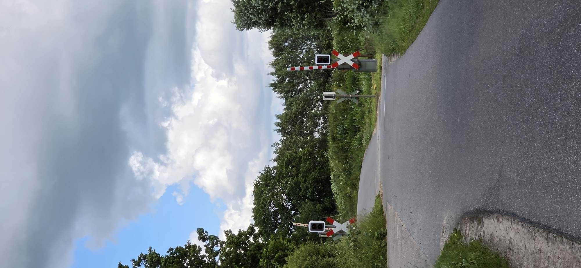 The image depicts a rural road in Maasbüll, Germany, leading towards a railway crossing. Two railway signals, each with a red and white striped pole and an "X" shaped crossing sign, are visible on either side of the road. The signal on the left has a black rectangular light above the crossing sign, while the signal on the right has a similar light and a red flashing light. The road is paved and appears to be in good condition, with grass and trees lining its sides. The sky is partly cloudy, with a mix of blue sky and grey clouds. There are no people or vehicles visible in the image.