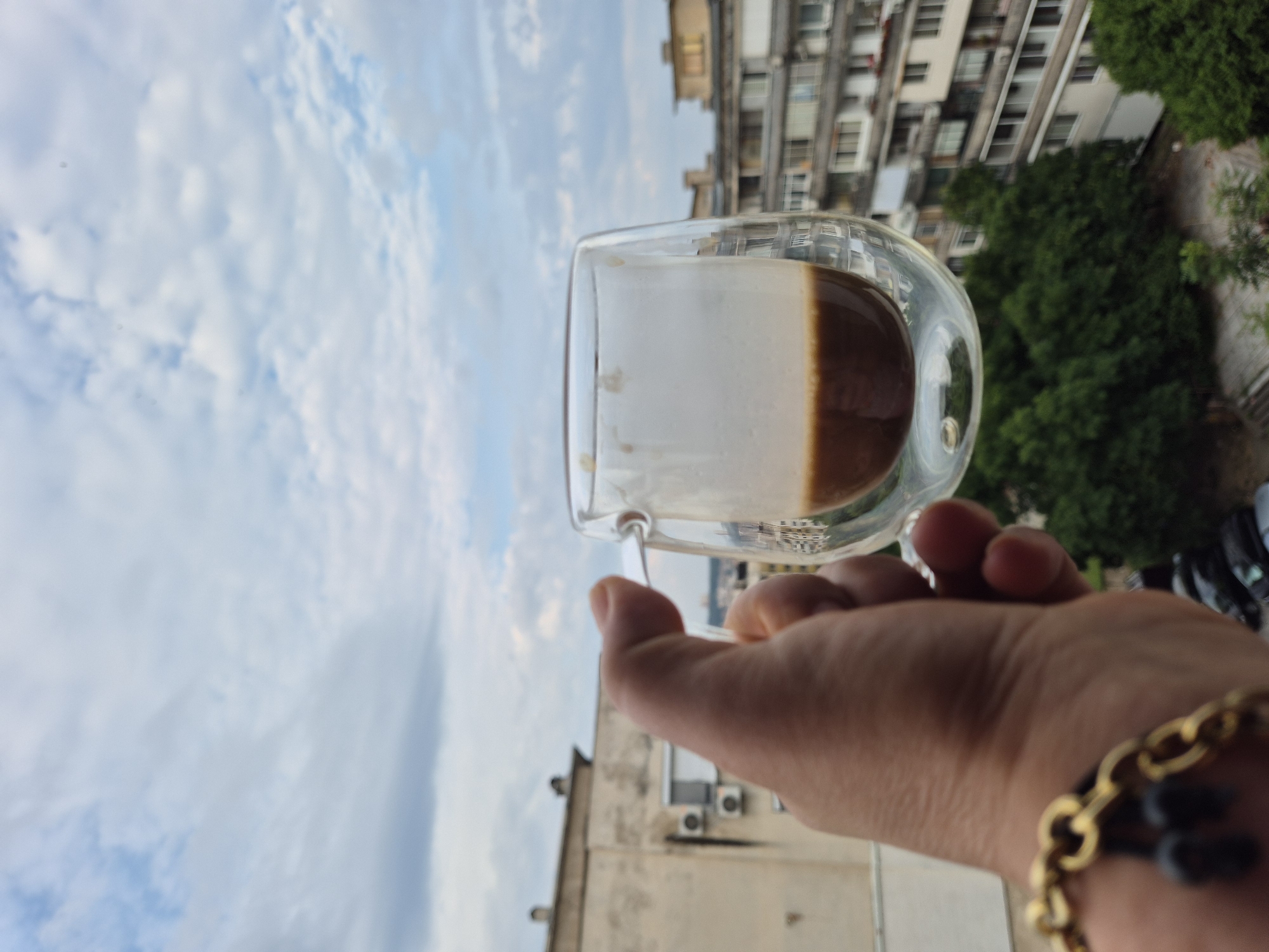 A hand wearing a gold bracelet holds a double-walled glass mug containing a dark beverage, possibly coffee, with a foamy top. The mug is held up against a cloudy sky. In the background, there are buildings, trees, and parked cars, indicating an urban setting. The perspective suggests the photo was taken from a balcony or window.