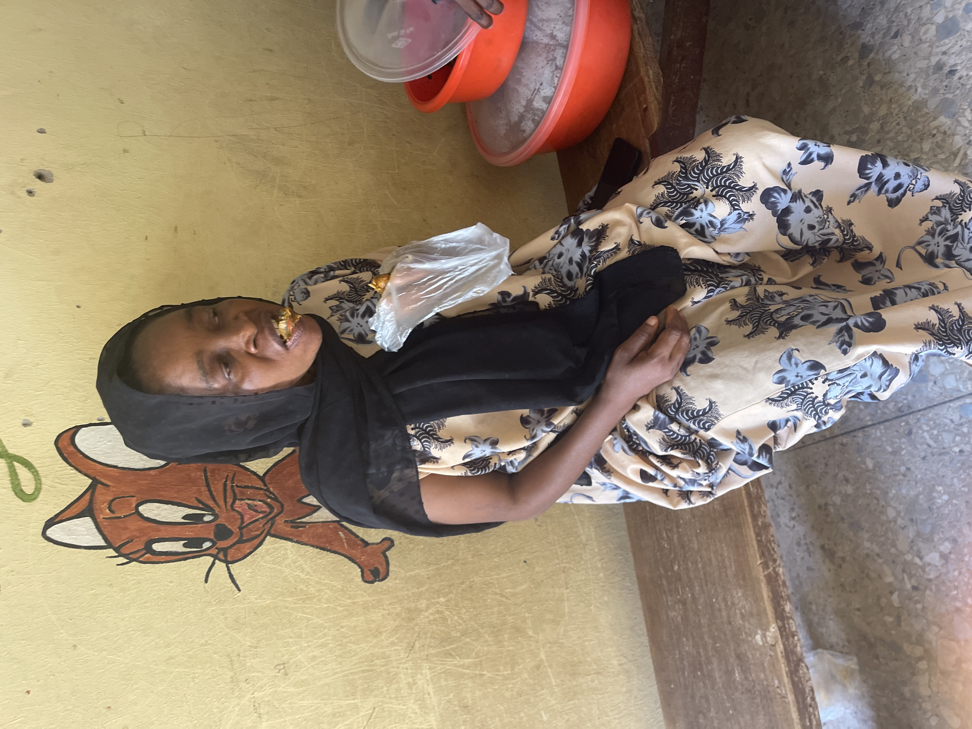 A woman is seated in an informal indoor or semi-indoor setting, smiling and holding a clear plastic bag containing a snack, which she appears to be eating. She is wearing a black hijab and a patterned dress in light beige or cream with dark floral designs. Her left hand rests on her lap.
To her left, a yellow wall features a brown and white cartoon drawing of a cat, resembling Tom from Tom and Jerry, along with a partial green drawing. The wall shows signs of wear. In the upper right part of the frame, two orange plastic basins are visible; one is partly covered with a clear lid, and the other contains a light-colored substance. A hand is partially seen near the top basin. The woman is likely sitting on a wooden bench or surface, part of which is visible on the right, and the floor appears to be concrete or tiled. No discernible text is visible.