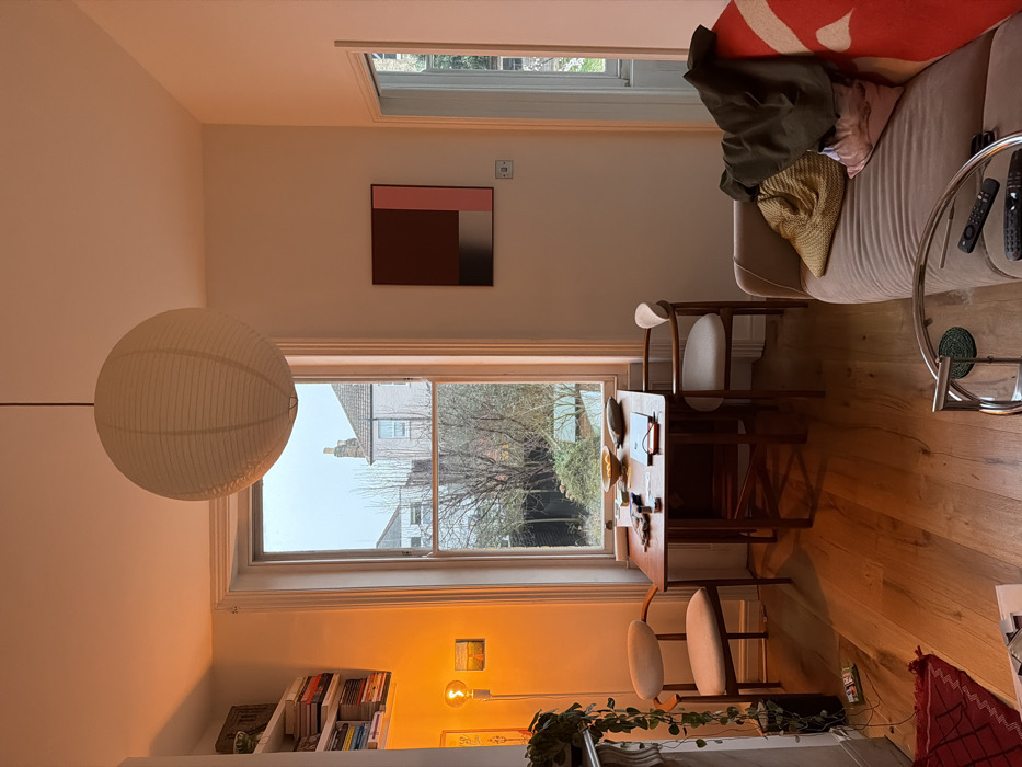 This image depicts a domestic interior, likely a living-dining area, located in a residential district of London, United Kingdom, evidenced by the characteristic brick architecture and sash windows visible through the main window. The room features light-colored walls and polished wooden floorboards.
A large, spherical white paper lantern hangs centrally from the ceiling. The dominant feature on the back wall is a tall, narrow sash window, offering a view of a garden with bare-branched trees and the rear facades of several traditional brick residential buildings. Directly in front of this window, a rectangular wooden drop-leaf dining table is positioned, flanked by two mid-century modern-style chairs with light upholstered seats. On the table, a yellow bowl containing several citrus fruits is visible, alongside a small notebook or tablet and other indeterminate small items.
To the left of the window, a white wall-mounted shelving unit holds multiple books. Below it, a section of a white fireplace mantelpiece is visible, on which a trailing green plant in a dark pot is placed. A tall, thin floor lamp with an exposed spherical incandescent bulb casts a warm orange glow onto this area, illuminating a small framed artwork on the wall nearby.
To the right of the window, a rectangular abstract artwork featuring blocks of dark red, light pink, and a gradient from dark to light grey is mounted on the wall. Further to the right, an open doorway leads to another room, where a window is also visible. In the foreground on the right, a beige fabric sofa is partially visible, draped with a dark green jacket, a straw-colored woven hat, and a red blanket with a large white stylized pattern. A round glass coffee table with a chrome frame is positioned in the immediate foreground, displaying several dark television remote controls. No individuals are present in the scene.