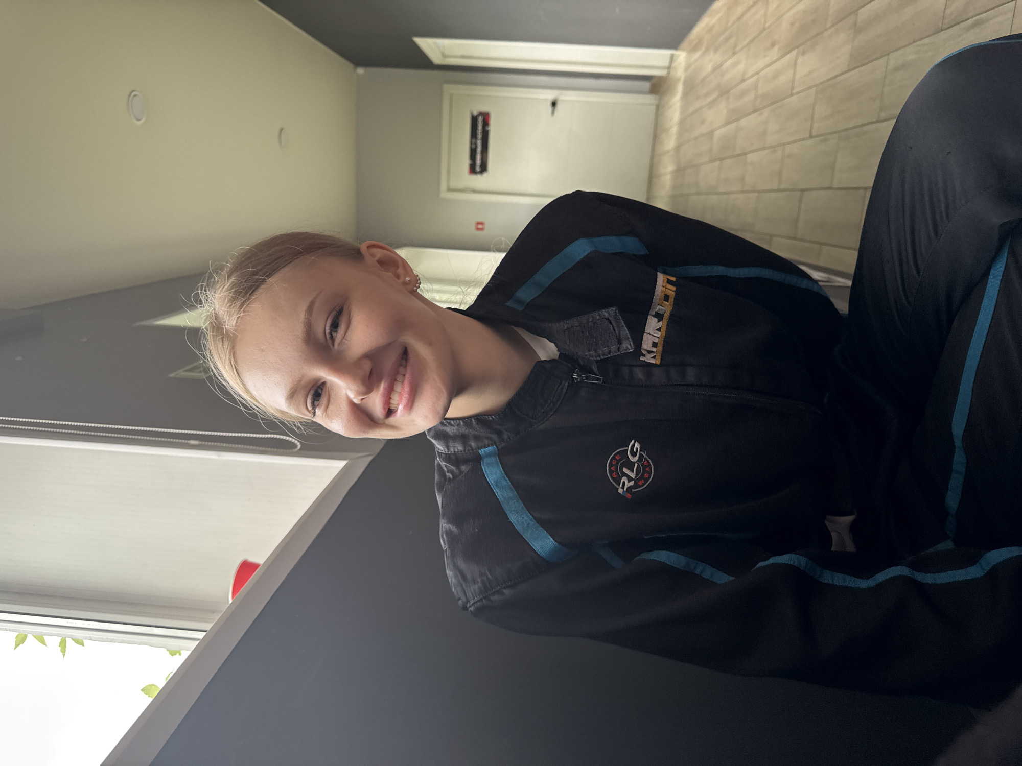 A fair-skinned woman with blonde hair is smiling at the camera. She is wearing a dark, zippered jumpsuit with blue stripes and the logo "RLG RACE WEAR". The background is an interior with light-colored walls on the left, and beige tiles on the right. A hallway with a door and a sign are visible in the background. The location is possibly a go-kart racing facility in Perm, Russia, given the jumpsuit and the hallway.