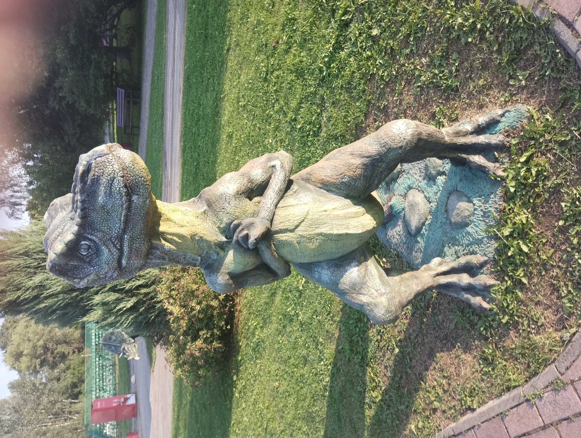 A full-shot, eye-level photograph captures a dinosaur statue positioned in an outdoor park setting. The statue, a dinosaur resembling a Tyrannosaurus Rex, is the central subject, depicted in a crouching stance with its arms held across its chest. The statue is rendered in shades of green and gray, with a textured surface simulating scales and skin. It stands on a rock-like base painted blue and green, surrounded by green grass and some patchy dirt.
In the background, the park features trees and bushes, indicative of a natural landscape. A paved walkway is visible on the left side of the frame, bordered by brickwork. Further in the background, a green fence and seating structures suggest the presence of spectator areas or picnic spots. The sunlight casts shadows, indicating it is daytime, likely late afternoon due to the warm lighting. The overall environment suggests a public park or an outdoor recreational area. The location is identified as the Sumy City Park in Sumy, Ukraine, a popular tourist destination known for its dinosaur-themed attractions.