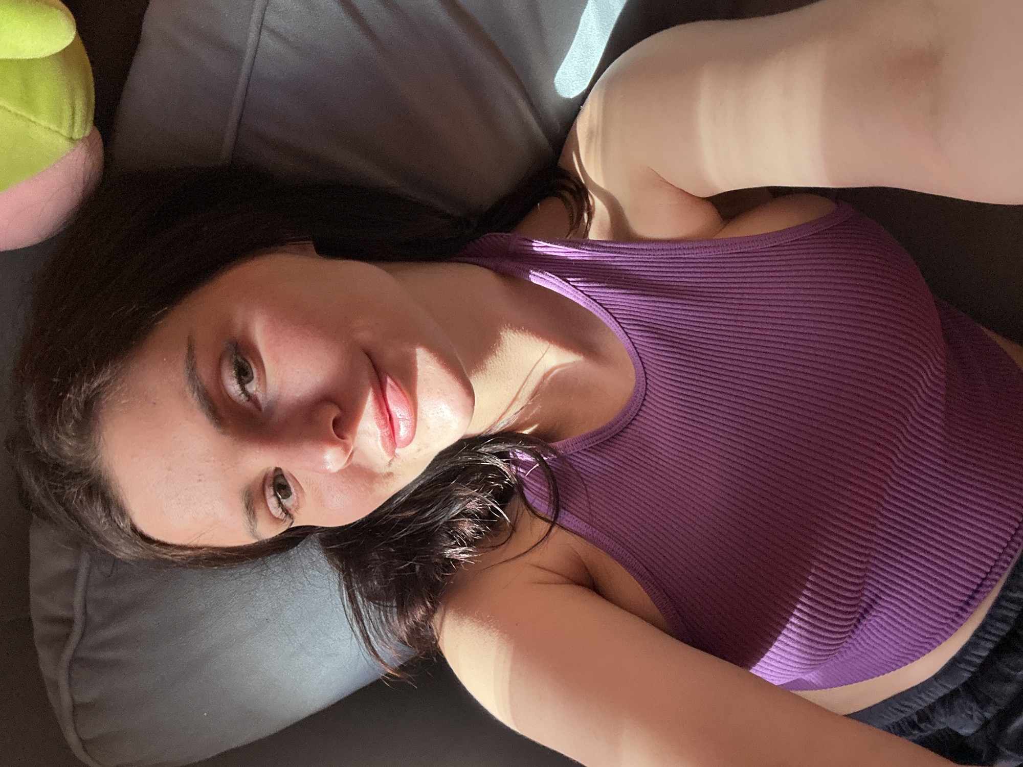 A young woman with dark hair lies on a gray couch, her body angled towards the camera. She wears a purple ribbed halter top and black bottoms. Sunlight streams across her face, highlighting her features, and casts shadows on her shoulder and chest. A soft green and pink object, possibly a stuffed toy, is visible in the upper right corner of the image, resting on the couch behind her. The background is dominated by the gray upholstery of the couch.
