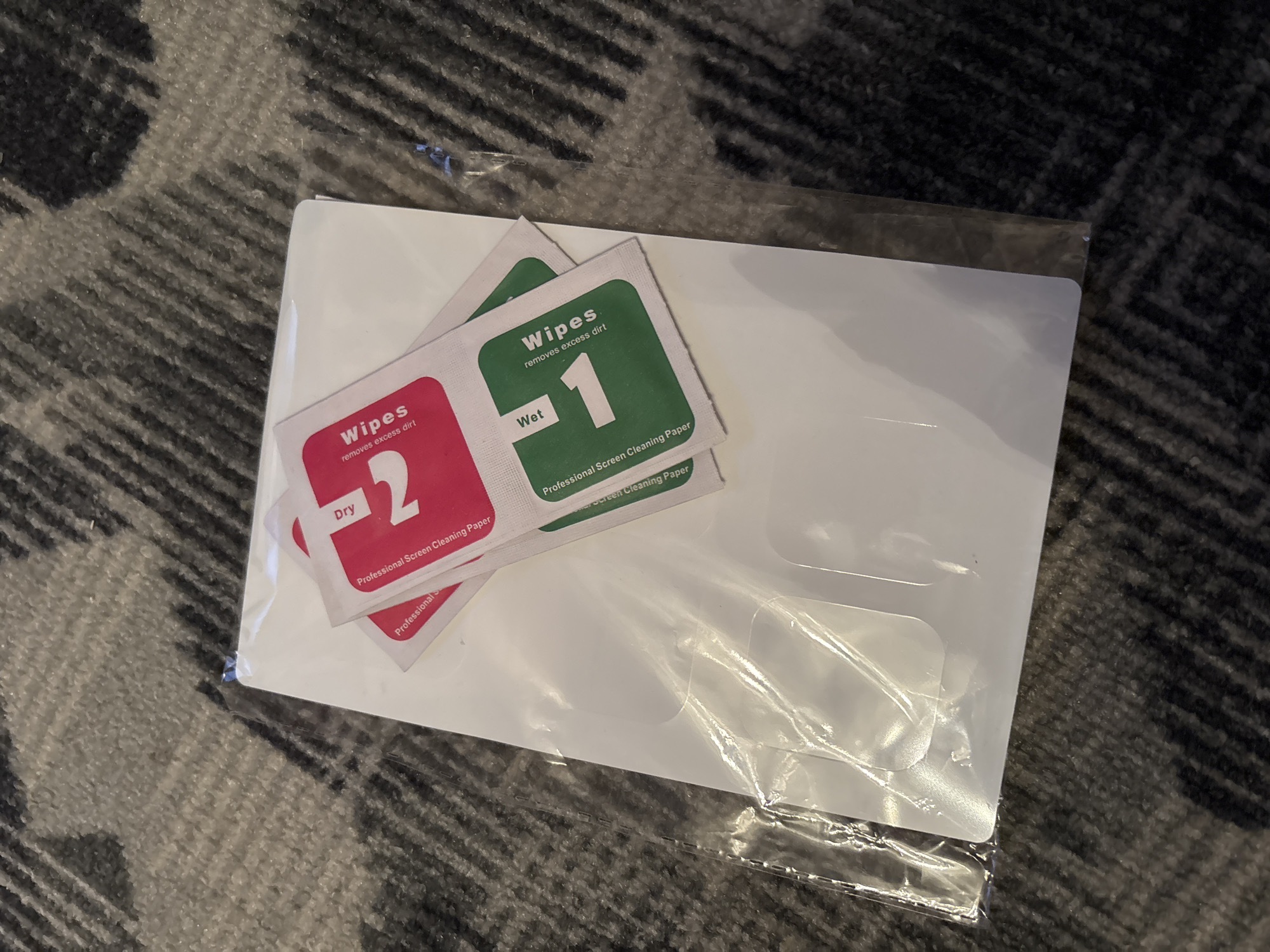 The image shows a set of screen protectors on a gray carpet. The screen protectors are white and rectangular, and they are still in their plastic packaging. There are also two small packets of wipes, one red and one green, which are also part of the screen protector kit. The red packet is labeled "Dry" and has a large number "2" on it. The green packet is labeled "Wet" and has a large number "1" on it. Both packets are labeled "Professional Screen Cleaning Paper" and "wipes removes excess dirt". The image is taken from a high angle, looking down at the screen protectors. The lighting is natural, and the focus is on the screen protectors. There is no indication of the location in Chernogorsk, Russia.