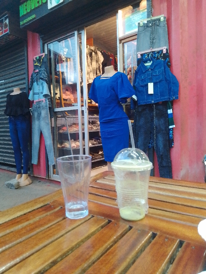 The image shows a wooden table with two empty glasses and a plastic cup with a green liquid, situated outside a clothing shop in Rungiri, Kenya (coordinates: -1.25, 36.684). The shop front is visible, displaying a variety of clothing items, including jeans, shirts, and a blue dress on a mannequin. The shop's facade features a red metal wall with a sign in the upper left corner. The floor is made of cement tiles. There are two mannequins outside the shop, both female, dressed in jeans and shirts. One of the mannequins is wearing a blue dress. The scene suggests a retail setting with a focus on clothing, where the table is likely used for customer seating or product display. The image captures a moment of quiet business activity, with no visible patrons. The light source appears to be coming from the shop windows, creating a slightly dim atmosphere.