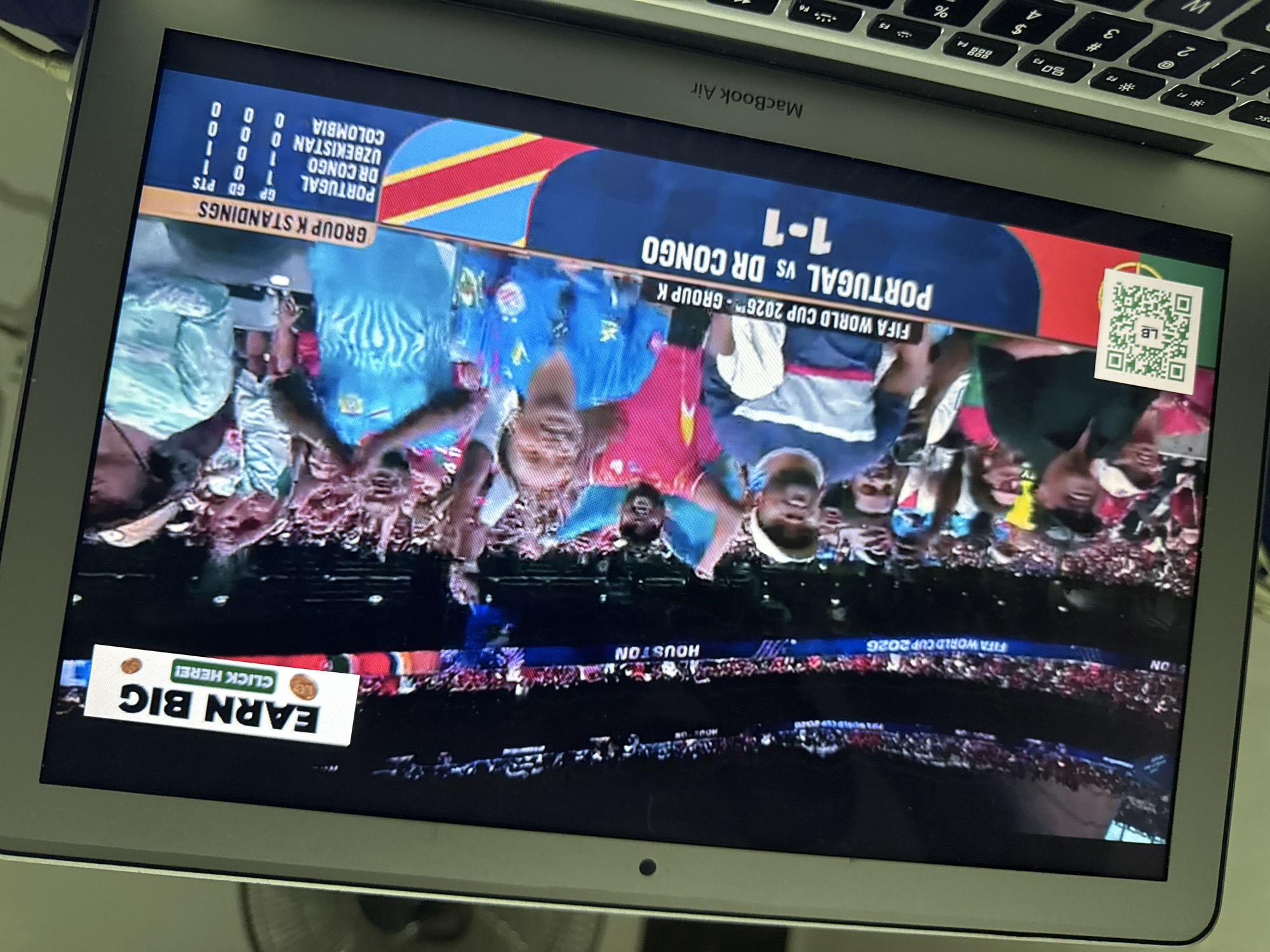 A laptop screen displays a live broadcast of a soccer match, with a score overlay indicating "PORTUGAL vs DR CONGO 1-1". The broadcast is for "FIFA WORLD CUP 2026 - GROUP K". In the foreground of the broadcast image, a crowd of enthusiastic spectators can be seen. One spectator in the front row is wearing a blue jersey. There is an advertisement banner at the top right of the screen reading "EARN BIG CLICK HERE!". A QR code is also visible on the left side of the screen. The broadcast also shows a "GROUP K STANDINGS" table. The laptop itself is open, with a keyboard visible at the bottom of the frame. The scene appears to be indoors, likely a room in Kufena, Nigeria, given the context.