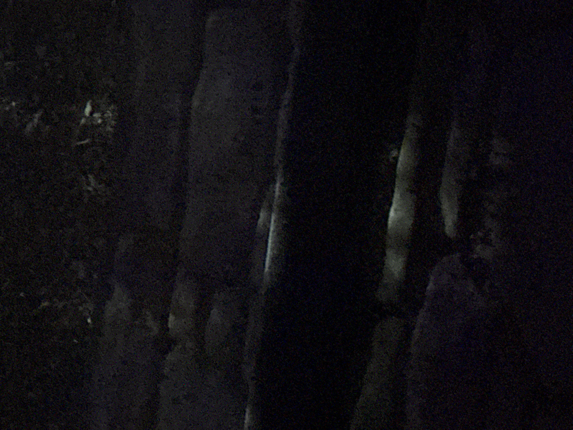 The image is a dark, grainy, aerial view of Vostok, Kyrgyzstan. The scene appears to be a nighttime landscape with some scattered lights. The terrain is uneven, with shadows obscuring many details. There are no discernible people, objects, or specific actions taking place. The environment is characterized by darkness and a rough, possibly rural, topography. The lighting suggests the presence of distant man-made illumination, but its source and purpose are unclear.