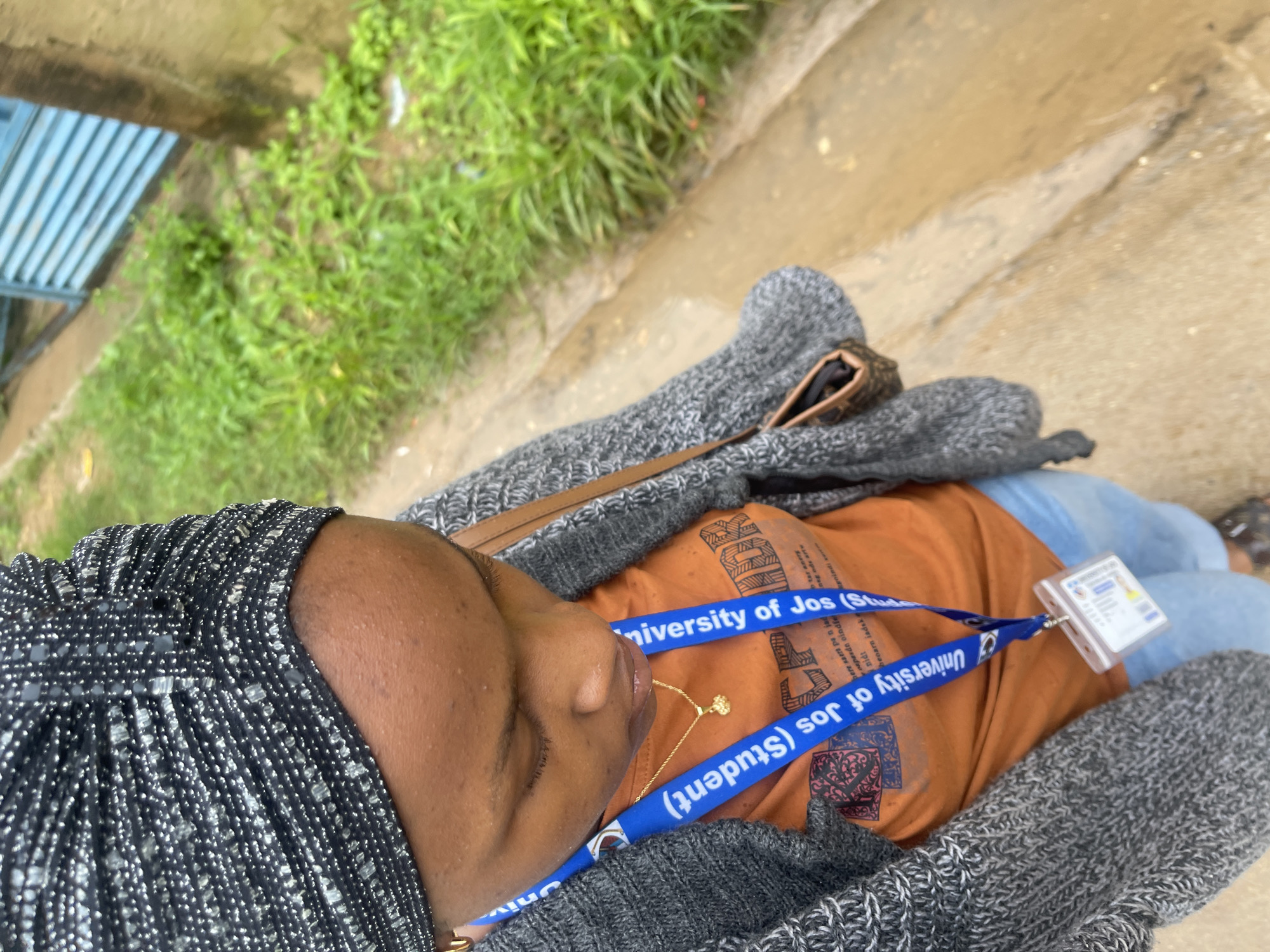 A woman wearing a grey knitted cardigan over an orange t-shirt and a black and silver patterned headwrap stands on a dirt path. She has a blue lanyard with a plastic ID holder and a gold necklace with a small charm. A small brown purse is slung over her shoulder. The background shows overgrown green grass, a dirt ditch, a concrete curb, and a portion of a blue metal fence. The scene appears to be in Rafin Par, Nigeria.