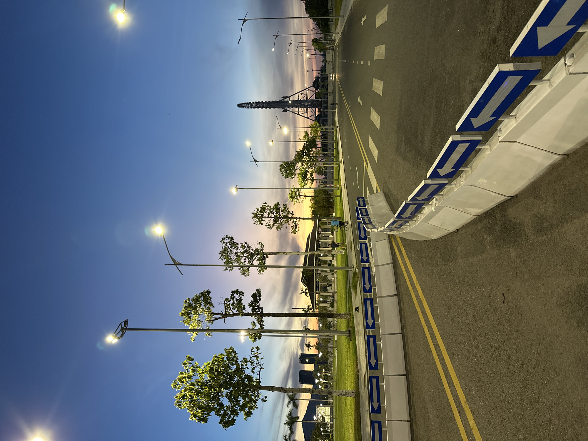 The image depicts a street scene at dusk in Đà Nẵng, Vietnam. A wide asphalt road with yellow dividing lines curves into the distance, marked with pedestrian crosswalks and white traffic barriers with blue directional arrows. On the left, a well-maintained green lawn is bordered by trees and palm trees. In the background, a pavilion-like structure with a dark roof stands under the twilight sky. Beyond that, two modern skyscrapers are visible, with more trees and streetlights scattered across the landscape. Dominating the far end of the road is a tall, skeletal Ferris wheel structure, silhouetted against the colorful sky. The sky itself transitions from a pale yellow and orange near the horizon to a deep blue overhead. Several streetlights illuminate the scene, casting a warm glow. Two individuals are present: one person is riding a bicycle on the road, and another is walking a bicycle on the sidewalk in the middle ground.