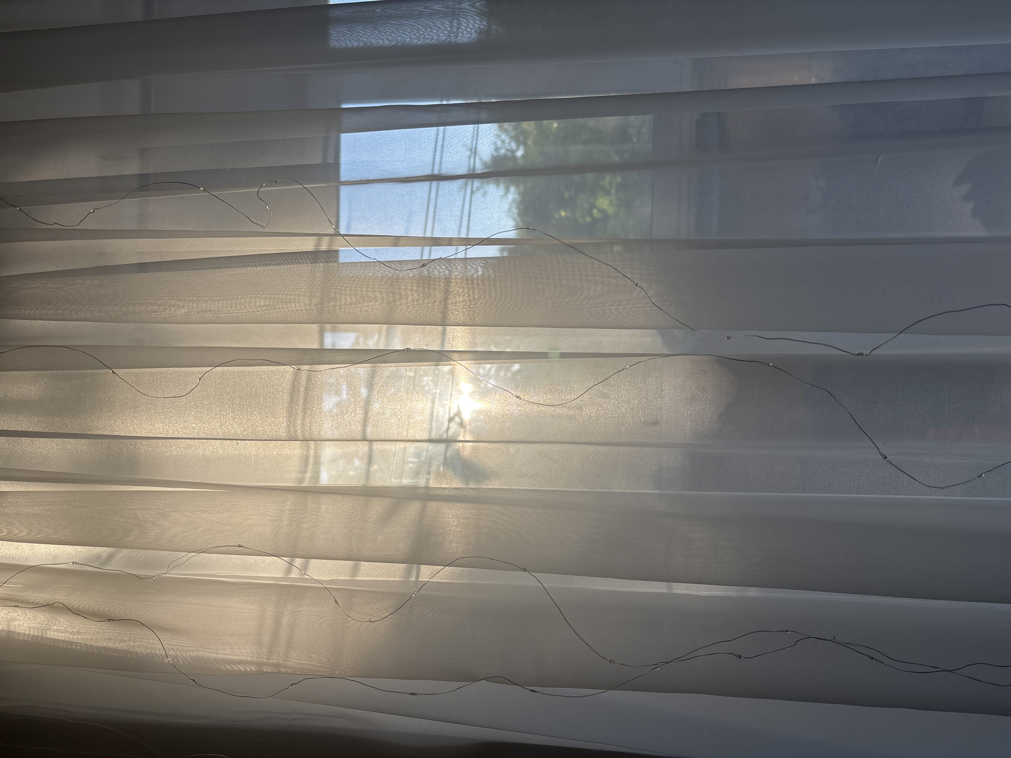 This is a full-frame, eye-level shot of sheer white curtains hanging vertically. Thin, wispy strings with small, round lights are draped across the curtains in wavy patterns. The sunlight is streaming through the window behind the curtains, creating a bright, diffused glow. The light is strongest in the center left of the image, casting a warm, golden hue. Through the translucent fabric, the silhouette of tree branches and power lines are visible against a bright blue sky on the right side of the image. Shadows of vertical window panes are also faintly visible on the curtains. The overall impression is one of soft, diffused light and gentle texture.