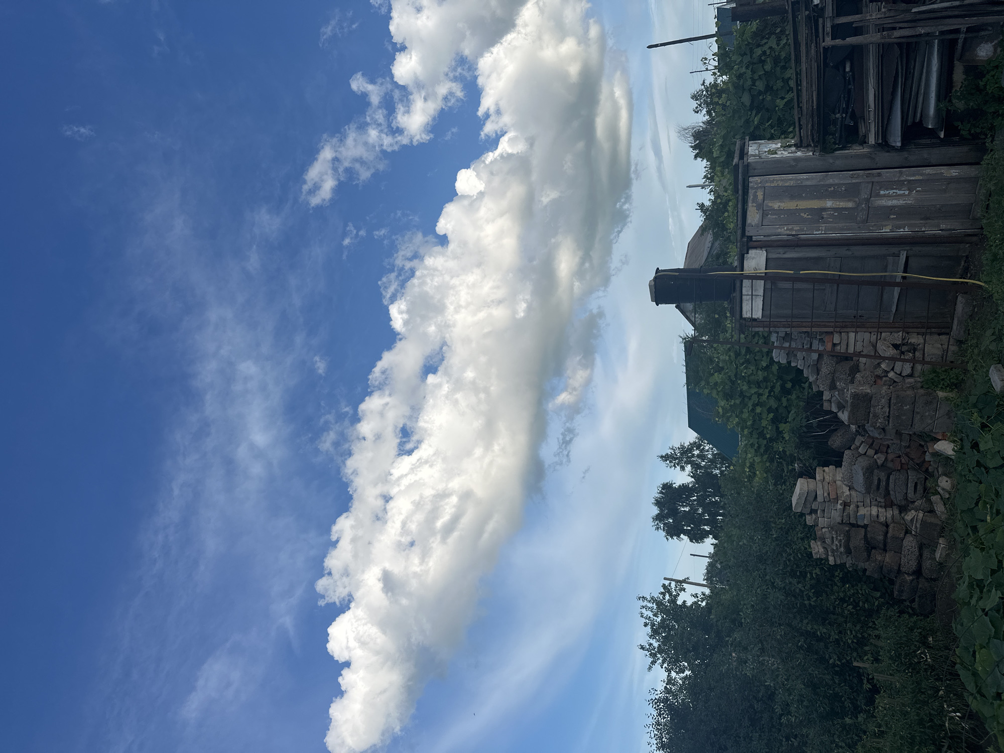 This media file captures a partly cloudy sky over a rustic scene in Tobyl, Kazakhstan. The main subjects are the sky and a collection of weathered wooden structures and materials.
The sky is a vibrant blue with distinct cloud formations. A large, billowy cloud stretches diagonally across the frame, casting shadows and highlighting its textured surface. Smaller, scattered white clouds dot the expanse. The overall impression of the sky suggests a bright day, likely late morning or early afternoon, with fair weather.
The ground level is dominated by an assortment of old, weathered wooden objects and building materials. These include what appear to be doors, planks, and possibly corrugated metal sheets, stacked and leaning against each other. Some of these items show peeling paint and signs of decay, indicating they are likely disused or part of a construction or demolition site. A pile of bricks is visible near the bottom right. Surrounding the structures are lush green trees and foliage, suggesting a rural or semi-rural setting. Power lines are also visible in the background, hinting at proximity to infrastructure.
There is no explicit activity or event taking place; the image appears to be a still-life composition of the environment. The scene evokes a sense of age and disuse, with the natural beauty of the sky contrasting with the man-made decay. The overall mood is calm and perhaps a little melancholic due to the state of the objects. No visible text is present in the image.