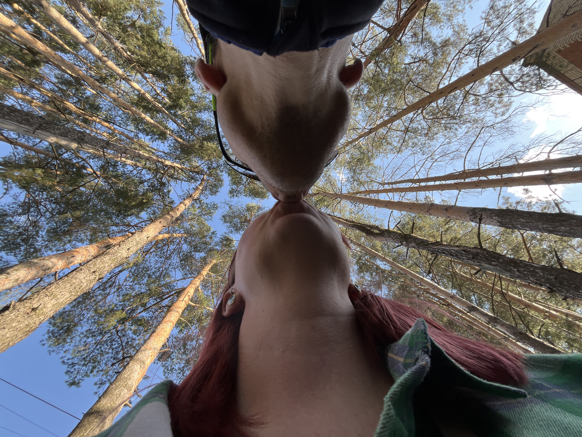 Captured from a low-angle perspective, the image features two individuals in close proximity with their lips touching, possibly kissing. Tall pine trees stretch upwards towards a blue sky with scattered clouds, framing the subjects. The person at the top of the frame has fair skin, visible ears, and a dark-colored collar. The person at the bottom has red hair, a green plaid shirt, and fair skin. The location is likely a wooded area within the city of Chaikovsky, Russia.