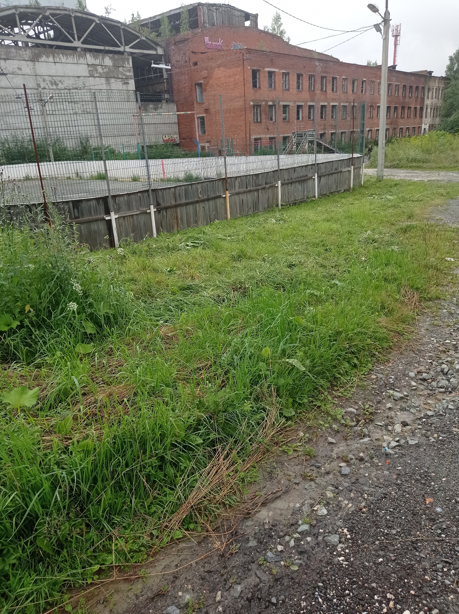 The image depicts an outdoor scene in Degtyarsk, Russia, focusing on an abandoned industrial complex. In the foreground, overgrown grass and weeds cover the ground, with some areas of mud and gravel. A weathered wooden fence runs horizontally across the mid-ground, separating the overgrown area from a fenced-off section with a wire mesh fence. Behind the fences are several dilapidated brick buildings, characteristic of industrial architecture, with broken windows and visible graffiti on some facades. A large, partially collapsed concrete structure with exposed metal framing is also visible. A street light pole stands to the right of the frame, near the brick buildings. The sky is overcast, suggesting a cloudy day. No people are clearly visible in the image.