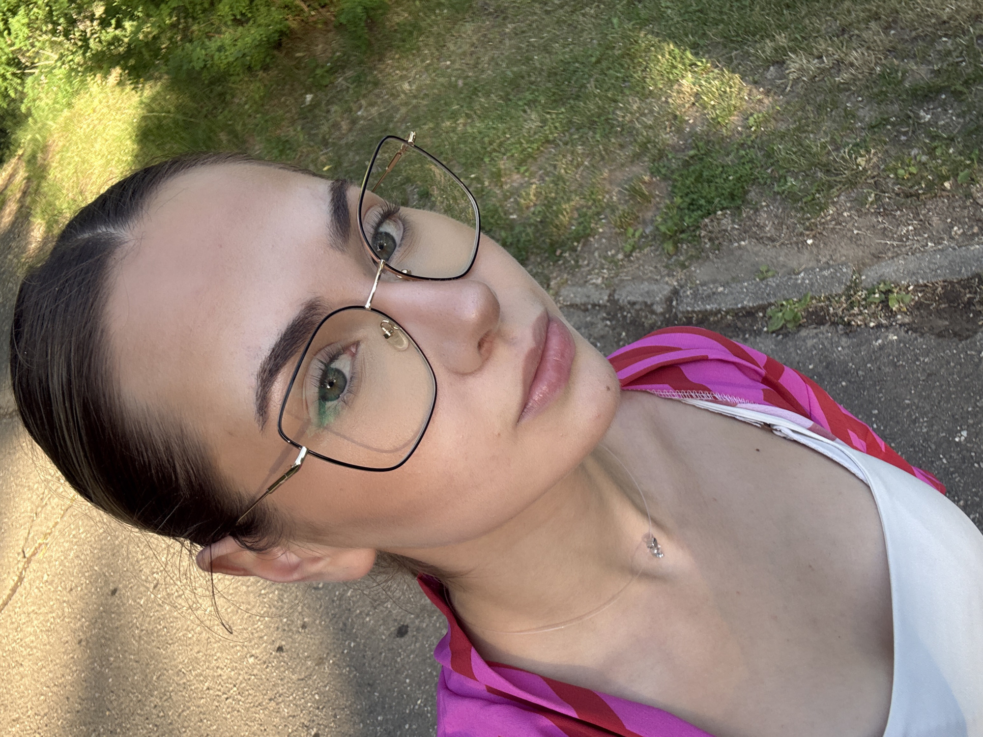 A woman with her dark brown hair pulled back and wearing black-framed glasses, a white top, and a pink and red patterned shirt is posing for a selfie. She is wearing a thin necklace with a small pendant. She is standing near a patch of grass and a gray concrete walkway. The photo appears to be taken outdoors in daylight. There is no specific information to indicate the location is in Blagoevgrad, Bulgaria.