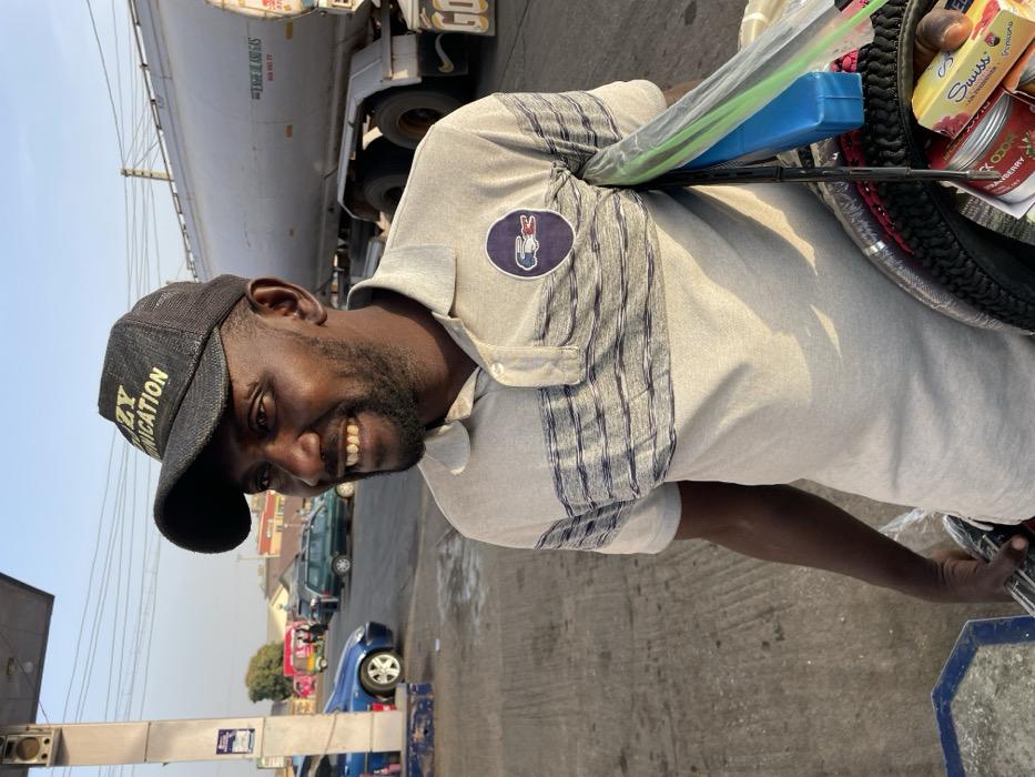 A man wearing a "Crazy Communication" baseball cap smiles while standing on a street. He has a short beard and wears a light-colored polo shirt with a patch on the chest. Behind him, a truck tanker is partially visible. He has a round container attached to his bike, filled with small items and packaged food. In the background, several cars are parked along the street, and power lines stretch across the sky.