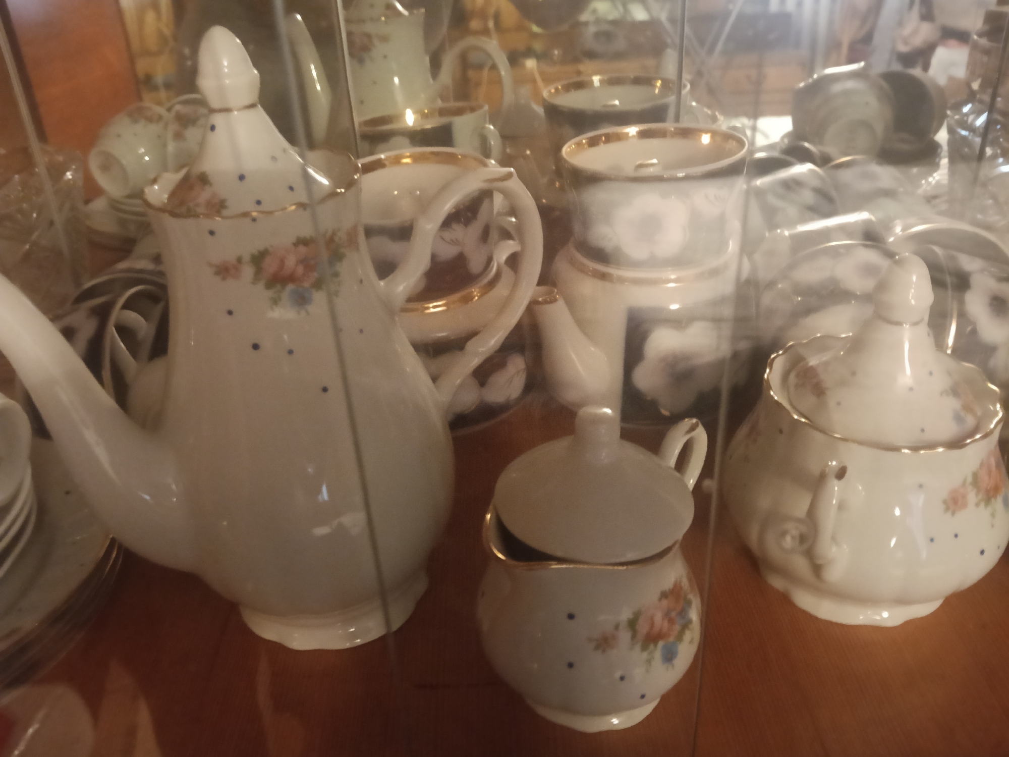 The image displays a collection of antique porcelain tea set items, including a large teapot, a creamer, and several cups and saucers, arranged on a wooden surface, likely within a display cabinet due to reflections. The porcelain is white with delicate blue polka dots and floral accents, rimmed with gold. The items appear to be well-preserved and are the primary focus of the photograph. The setting is a domestic interior in Rybnitsa, Moldova. There are no people visible in the image. The lighting is dim, contributing to a slightly hazy quality and the prominence of reflections.