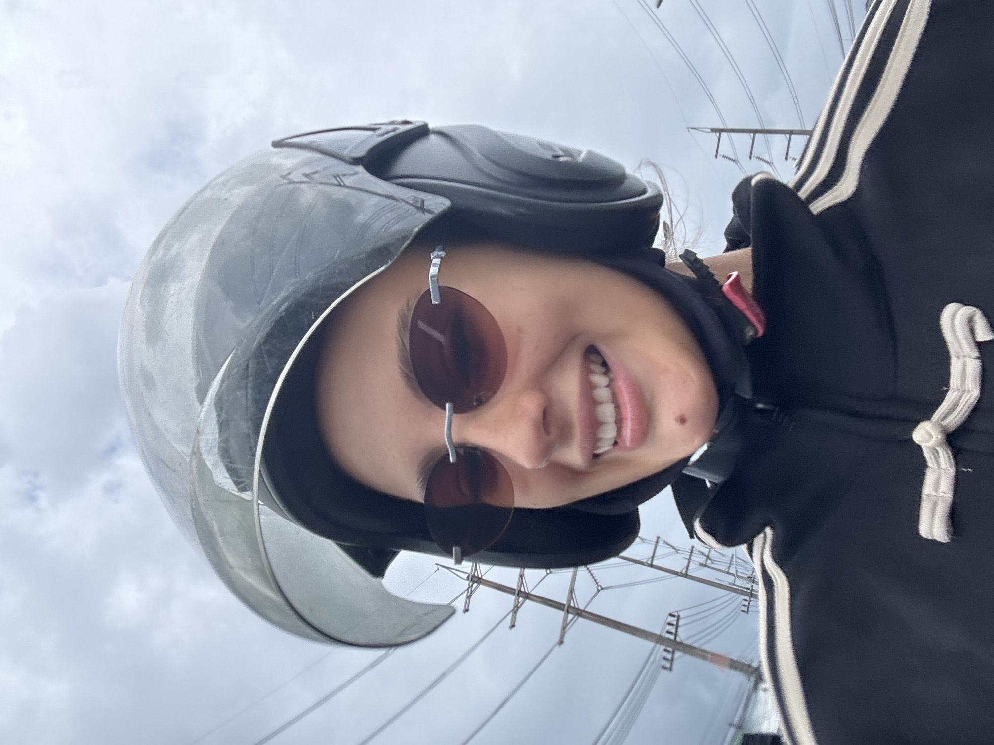 A female subject, wearing a black motorcycle helmet with a partially opened visor and brown round sunglasses, is smiling. She is also wearing a black jacket with white stripes on the sleeves. Power lines are visible in the background. The scene appears to be in Wichit, Thailand.