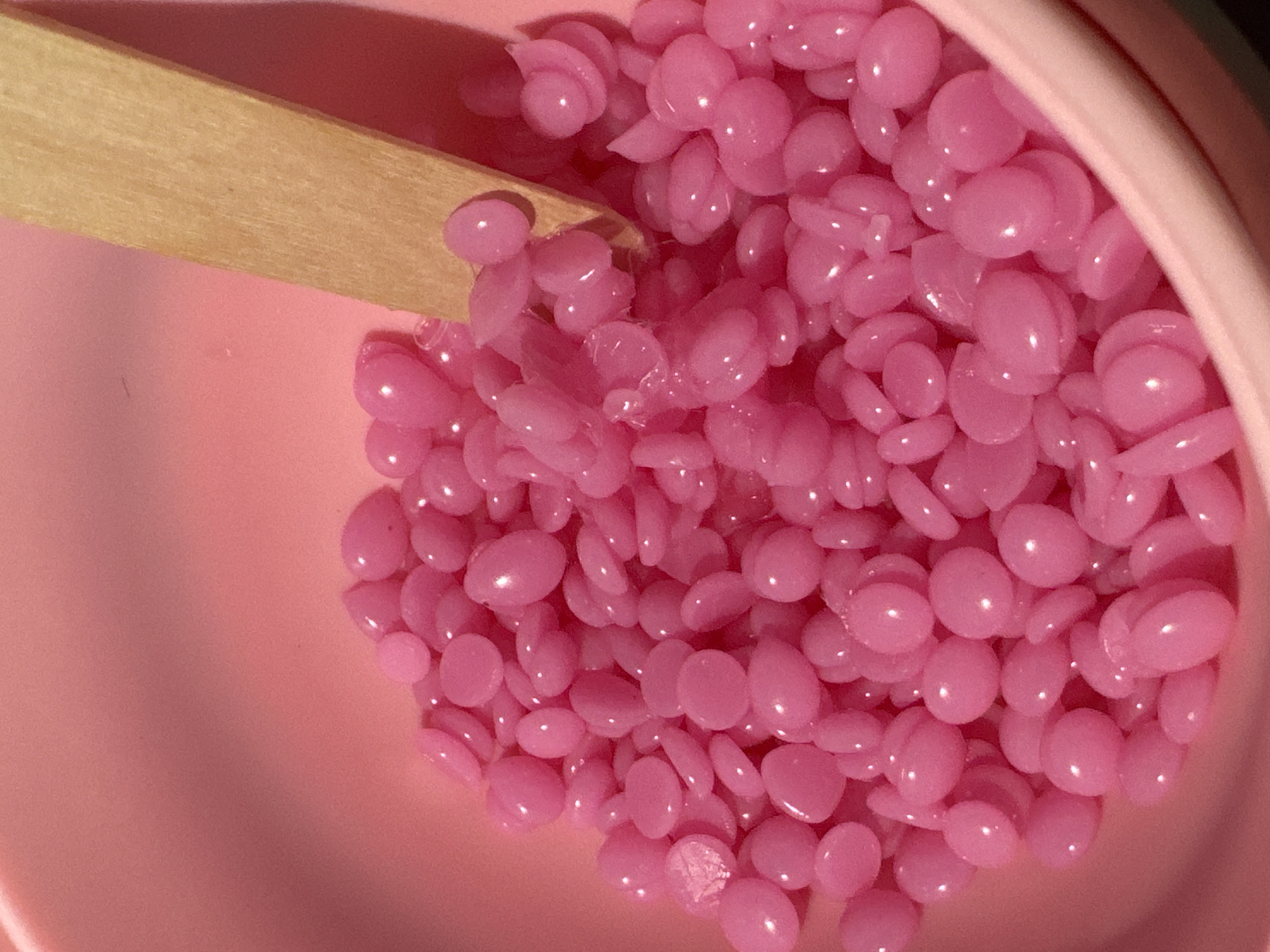 A close-up, high-angle shot shows a pink bowl filled with small, pink, pearl-shaped beads. A light-colored wooden stick is positioned diagonally across the top right of the bowl, partially submerged in the beads. The lighting casts shadows on the beads and the bowl, highlighting their glossy texture. The background is a soft pink, consistent with the color of the bowl. The image is a still life of a cosmetic product, likely wax beads for hair removal, and a wooden applicator.