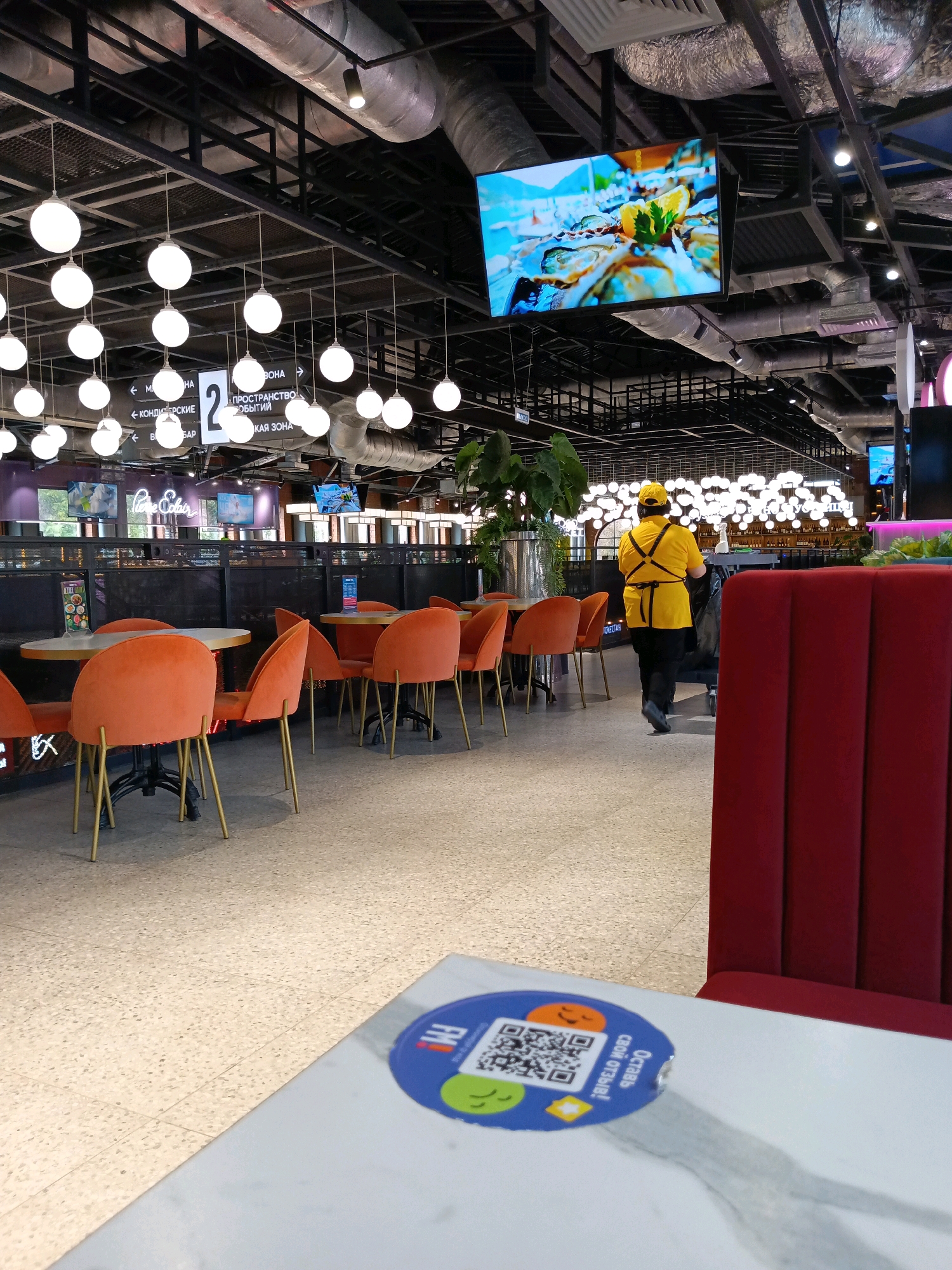 The image depicts the interior of a modern, brightly lit food court or restaurant with a capacity for several patrons. The setting features a high ceiling with exposed ventilation systems and numerous circular pendant lights. The floor is tiled with a speckled pattern. Orange chairs with gold legs are arranged around white tables, and a plush red seating area is visible on the right. A large screen displaying a close-up image of oysters and lemon hangs from the ceiling. A person wearing a bright yellow uniform, including a cap and apron, is walking away from the camera, pushing a cleaning cart. On a table in the foreground, a blue and orange circular sticker with a QR code and playful emojis is visible. This sticker appears to be a call to action for customers to leave reviews. The food court has directional signage, including a sign with the number "2" and text in Russian indicating different zones like "Confectionery" and "Activity Space." The overall atmosphere is one of a commercial establishment designed for dining and relaxation. The presence of Cyrillic text on the signage strongly suggests the location is in Russia. Given the architectural style, lighting, and general ambiance, this scene is likely located within the OZ Mall in Krasnodar, Russia, a popular shopping and entertainment complex known for its diverse food court offerings.